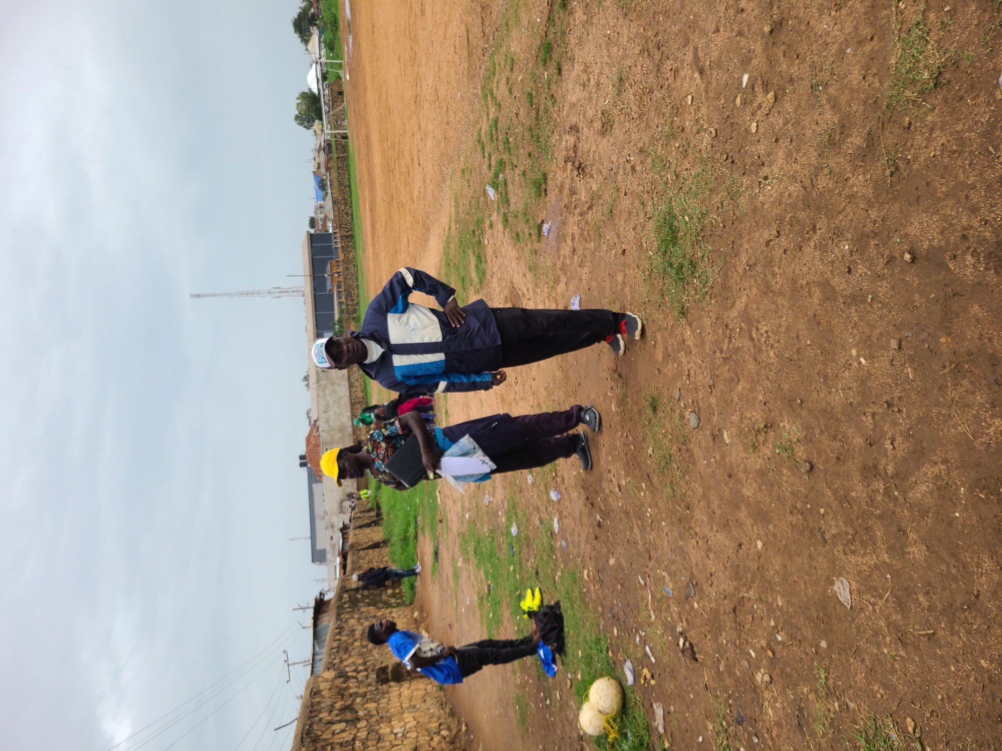 Three men are standing on a dirt field next to a stone wall. The first man is wearing a blue and white jacket, black pants, and a white hat. He has his hands on his hips. The second man is wearing a yellow hat, a colorful shirt, and black pants. He is holding a laptop. The third man is wearing a blue shirt and black pants. There are two soccer balls near him.