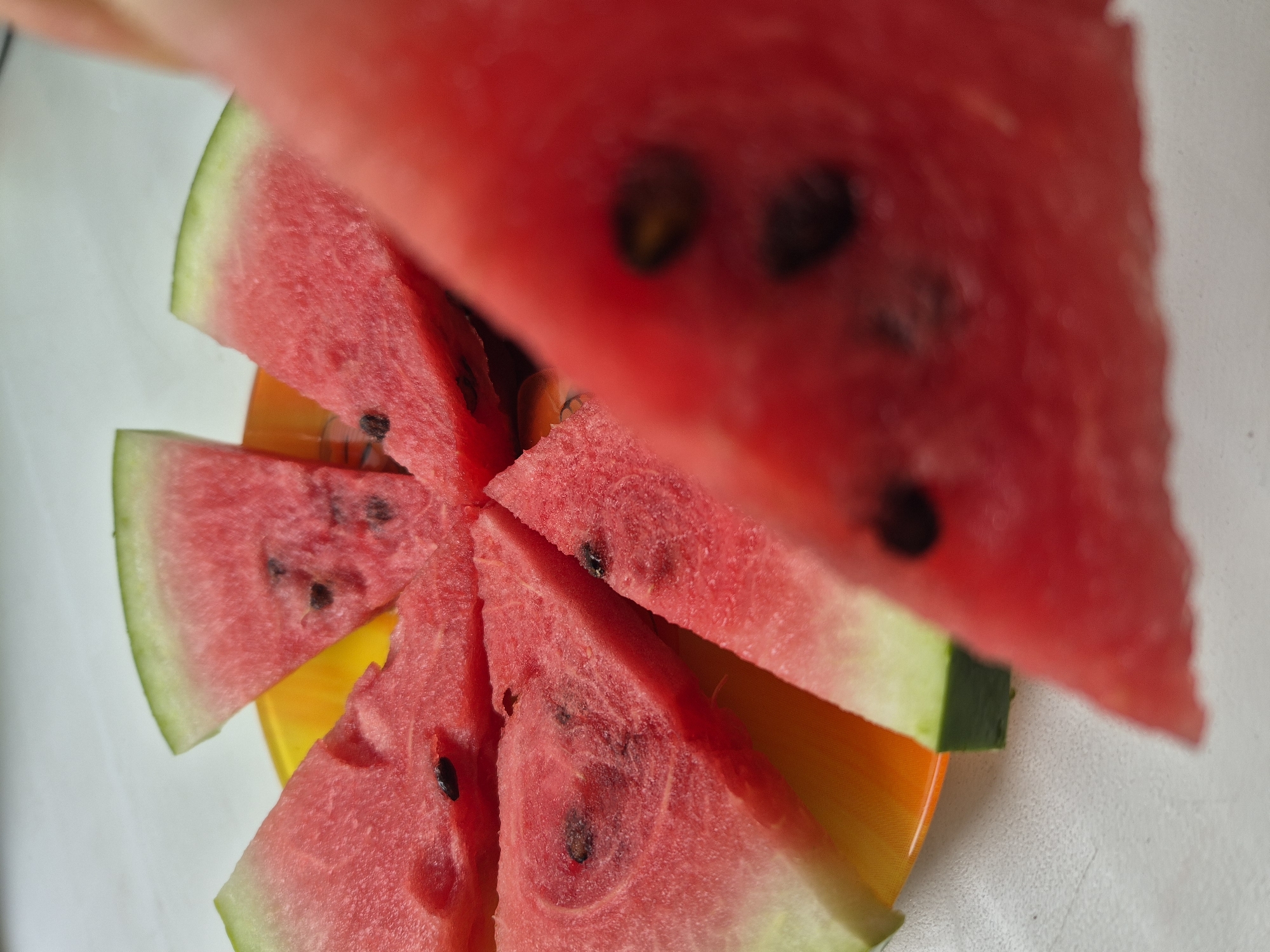 The image shows several slices of watermelon arranged on an orange plate. A large slice of watermelon is in the foreground, out of focus. The background is a white surface, possibly a table. The scene is not located in Kokshetau, Kazakhstan.