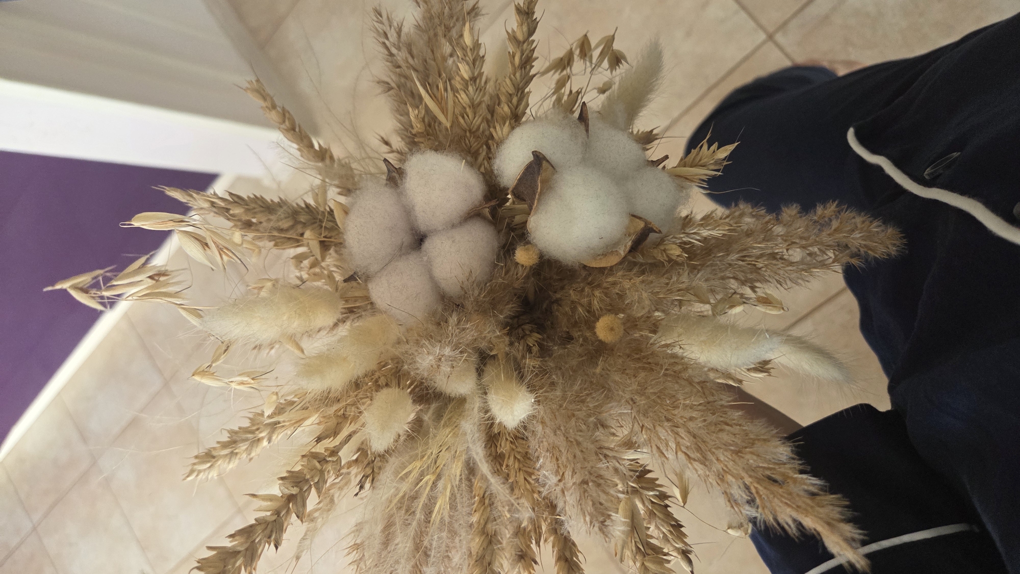 A bouquet of dried flowers, including wheat, bunny tails and cotton, is held in someone's hand. The person is wearing a navy blue top with a white stripe. The background includes a light-colored tiled floor and a light purple wall. The location is not specified, and cannot be discerned from the image.