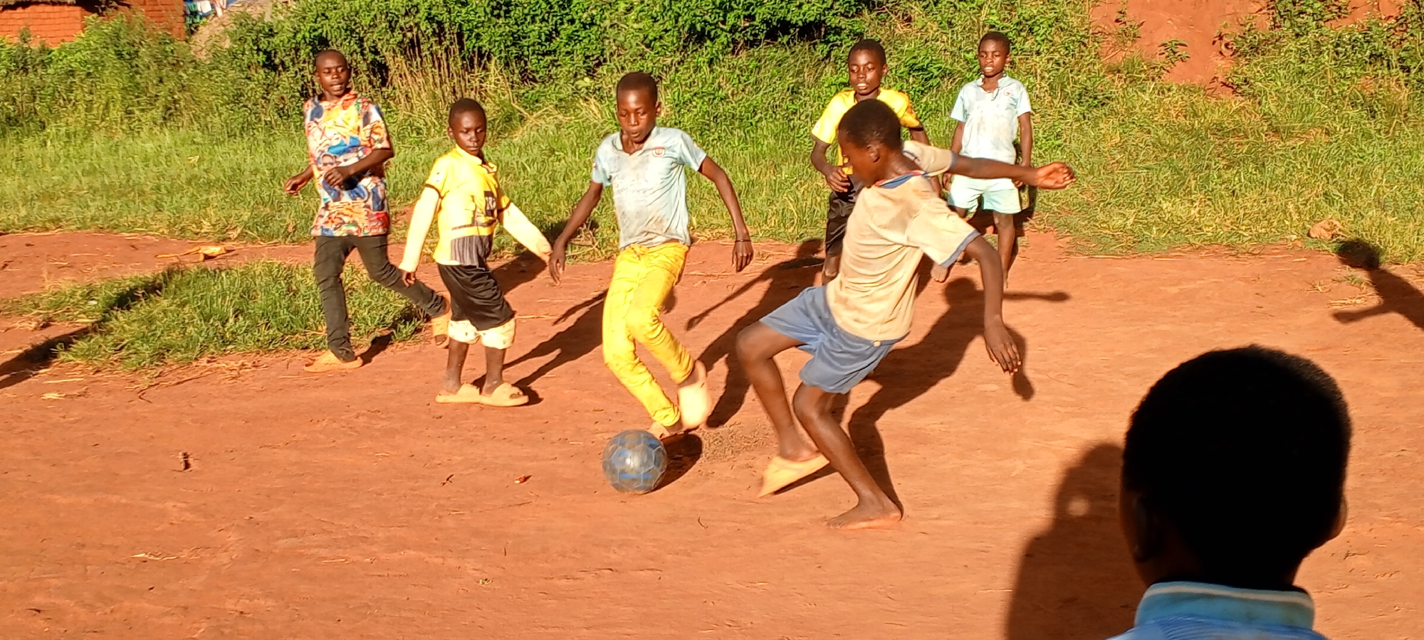 In Ngalula, Democratic Republic of the Congo, a group of six young boys play soccer on a red dirt surface. One boy, barefoot and wearing blue shorts and a light-colored t-shirt, kicks a blue soccer ball. Another boy in yellow pants and a light blue t-shirt approaches. A boy in a yellow and black outfit and sandals stands nearby. Another boy wearing a shirt with a graphic print and black pants is also present. Two other boys in light-colored shirts stand in the background. The scene is set in a dirt field with green foliage in the background.