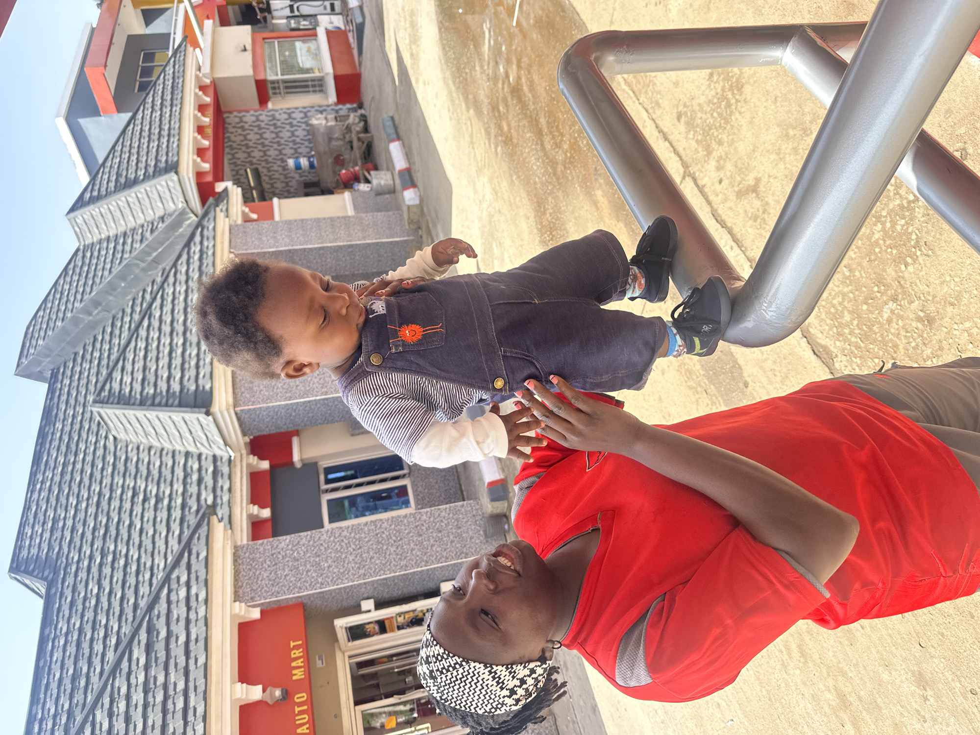 A Black woman with a patterned headscarf and a red shirt stands outdoors, looking at a young Black child who is wearing a striped shirt and dark blue overalls. The child is standing on a silver railing, looking down. The woman is holding the child's hand, seemingly guiding or supporting them. In the background, a building with red trim and a dark grey roof is visible, with signage indicating "AUTO MART". The ground appears to be paved, with some areas wet or reflective, suggesting recent rain or cleaning. The scene appears to be in a commercial area, possibly a gas station or shopping center. The sky is clear and blue, indicating daylight. The overall lighting suggests a sunny day.