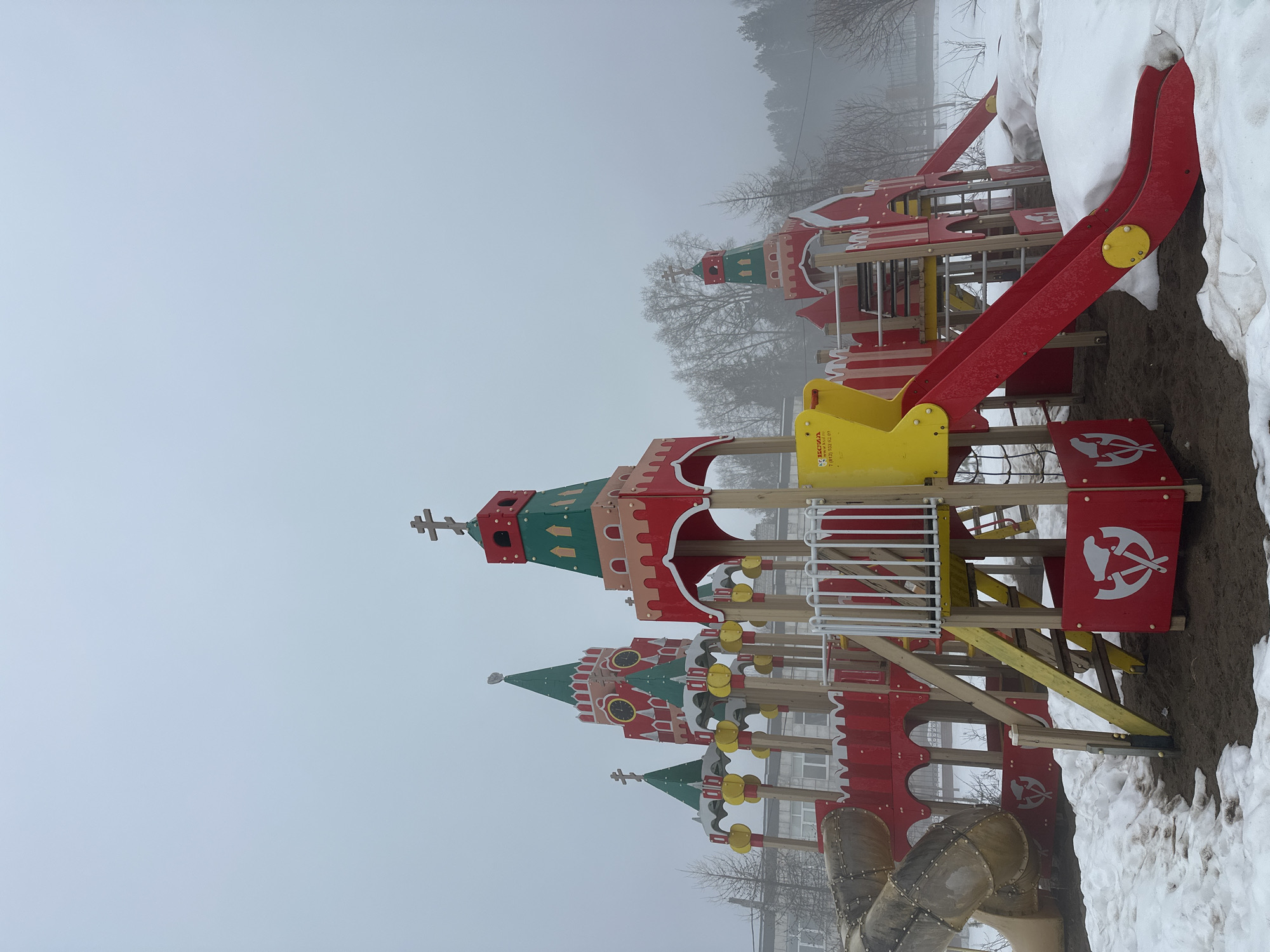 A playground structure in the style of the Moscow Kremlin towers is depicted in the city of Chaikovsky, Russia. The structure is painted in red, green, and yellow. It includes slides, climbing frames, and stylized crenellations. The ground is covered in snow and dirt, and the sky is overcast. Bare trees are visible in the background, obscured by fog. No people are visible.