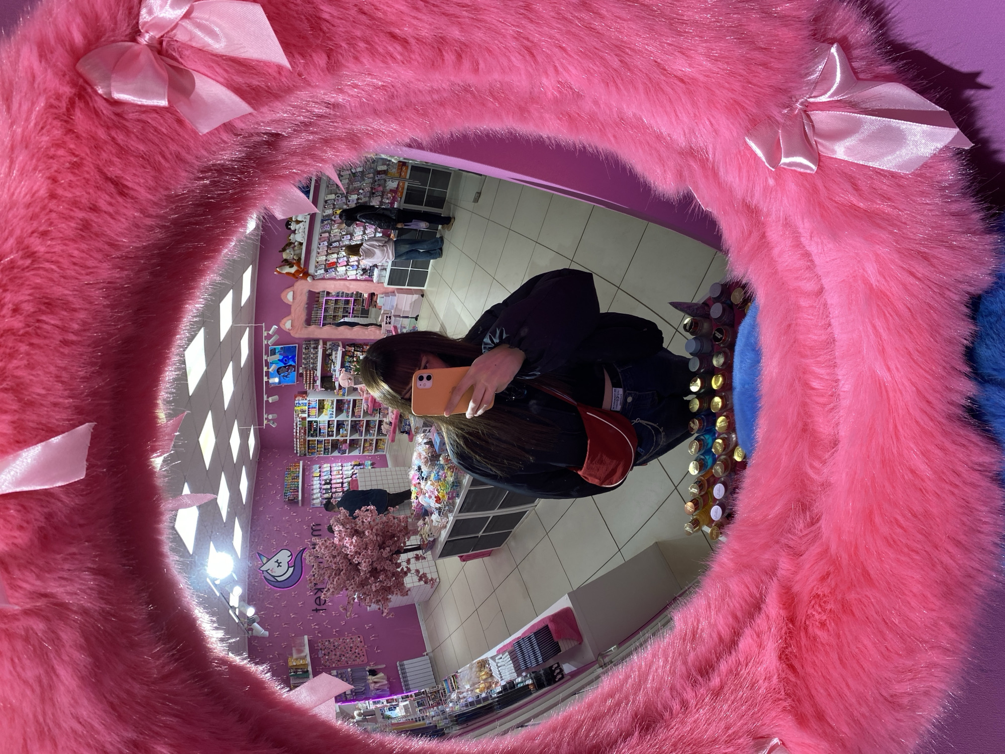 A person reflected in a circular mirror with a pink faux fur frame and pink ribbon bows is holding a smartphone, presumably taking a selfie. The mirror is reflecting the interior of a retail store with pink decor. Store shelves stocked with merchandise can be seen in the background, along with other customers. The location appears to be a shop with a distinctive pink color scheme and a unicorn logo, possibly a store in Votkinsk, Russia, based on the prompt. The mirror is the main subject of the image.