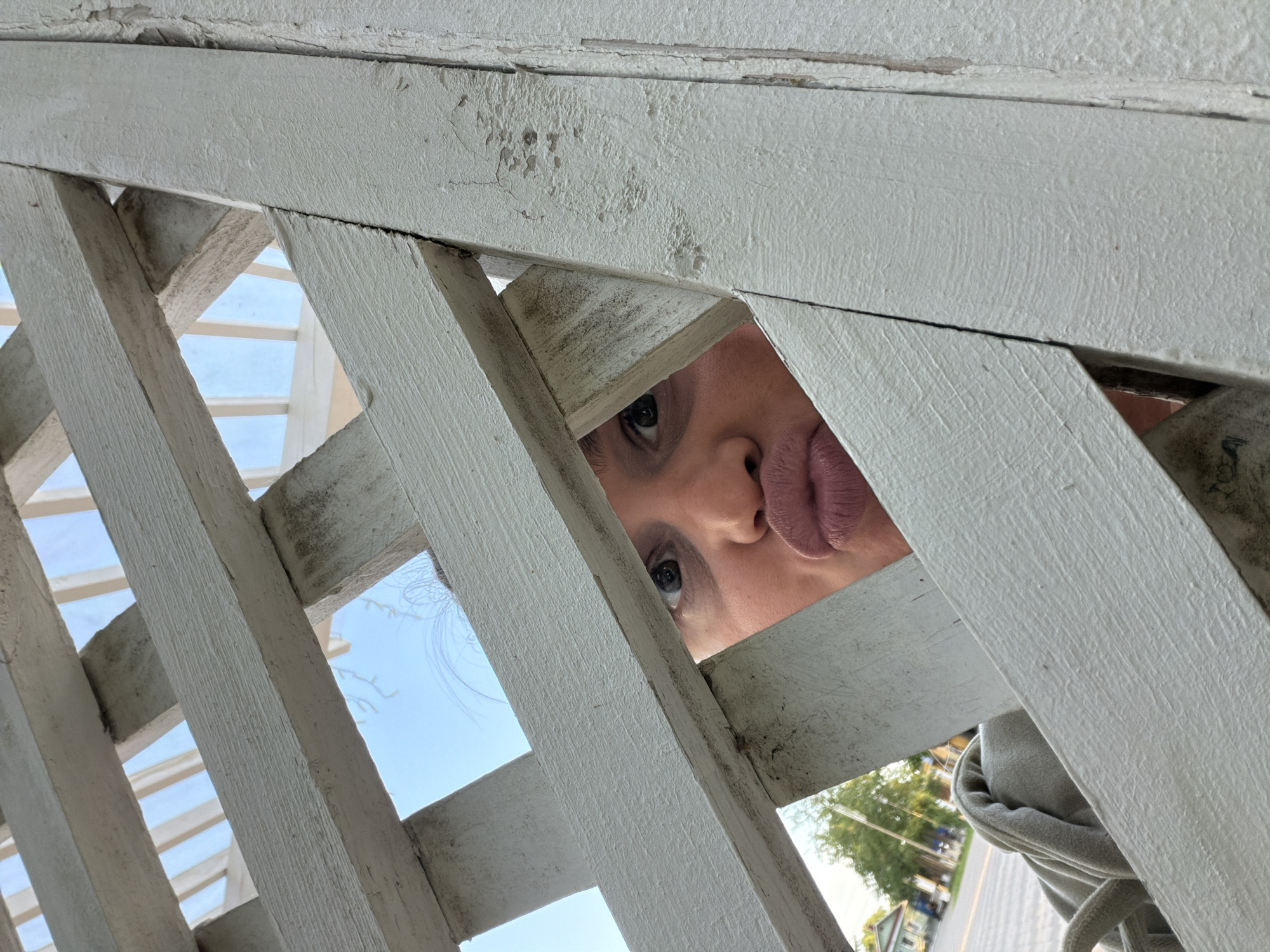 The image shows a person with their face pressed against a white wooden lattice. The person has light skin, dark eyes, and pursed lips. The lattice is aged and the paint is chipped in some areas. Through the lattice, one can see a blue sky, a few buildings, and trees, suggesting an outdoor urban environment. The trees are green and lush with a street in the background. While the request specifies Sarapul, Russia, there is nothing in the image to confirm this location forensically.