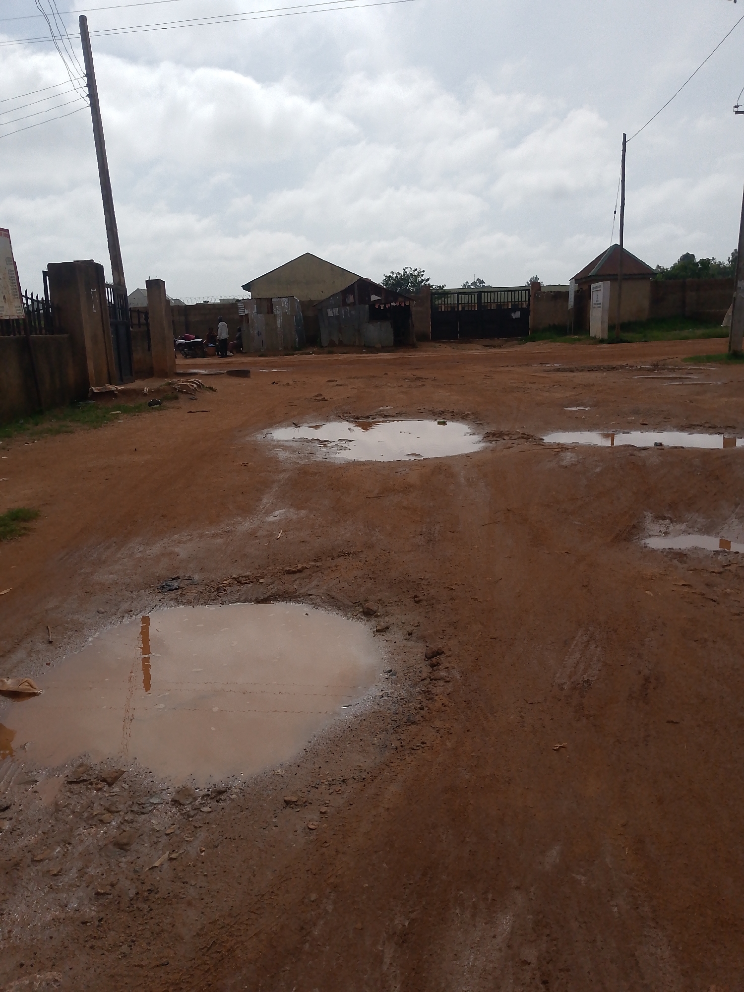 The image depicts an outdoor scene in what appears to be Barakin Angundi, Nigeria. The foreground is dominated by a dirt road with several large puddles of muddy water, indicating recent rainfall. Some of these puddles reflect the cloudy sky and nearby objects. In the middle ground, there are several structures. To the left, a large concrete pillar and a metal gate are visible, with a building behind them. Further back, a building with a pitched roof and a smaller structure with a peaked roof and a sign are present, situated near a large black metal gate. A few individuals are standing near a motorcycle and some buildings in the mid-ground. The background shows more buildings and trees under an overcast sky. Electrical poles and wires are visible in the upper part of the image. The overall impression is of a rural or semi-urban area that has experienced wet weather.