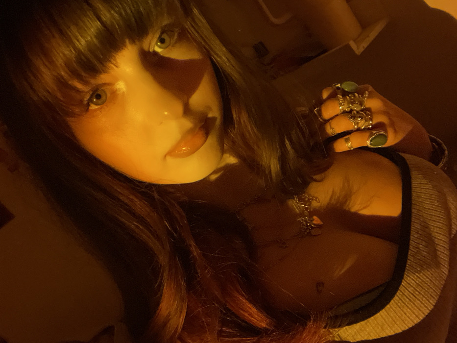 A woman with bangs poses in a low-lit, close-up shot. Her skin is fair and illuminated by a warm, yellow glow. She has striking eyes. She is wearing multiple rings on her hand and a necklace adorns her chest. The collar of her shirt is visible, showing a light-colored material with a dark trim. The style of the photo suggests a candid, almost intimate portrayal.