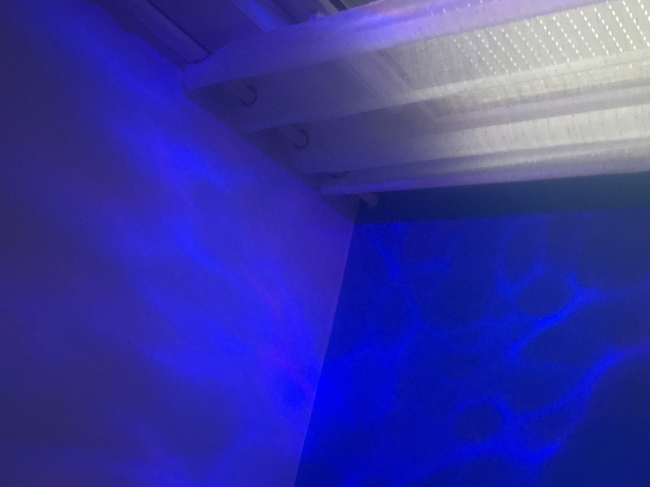 This image depicts an indoor scene within a structure located in Conflans-Sainte-Honorine, France. The environment is a room illuminated by artificial blue light, casting dynamic, ripple-like patterns onto the surfaces. The upper portion of the frame shows a light-colored ceiling, illuminated with undulating blue light. Below the ceiling, a wall, appearing to be a solid, darker color (possibly blue or a dark neutral shade), is also covered with prominent blue light patterns resembling reflections from water. To the right, a section of a window is visible, partially obscured by a sheer, light-colored curtain, likely white or off-white. This curtain features grommet-style rings at its top, through which it is suspended from an unseen rod or track system. The curtain material exhibits a patterned texture, possibly composed of small, raised dots or a grid. No individuals, animate subjects, or specific actions are depicted within the frame.