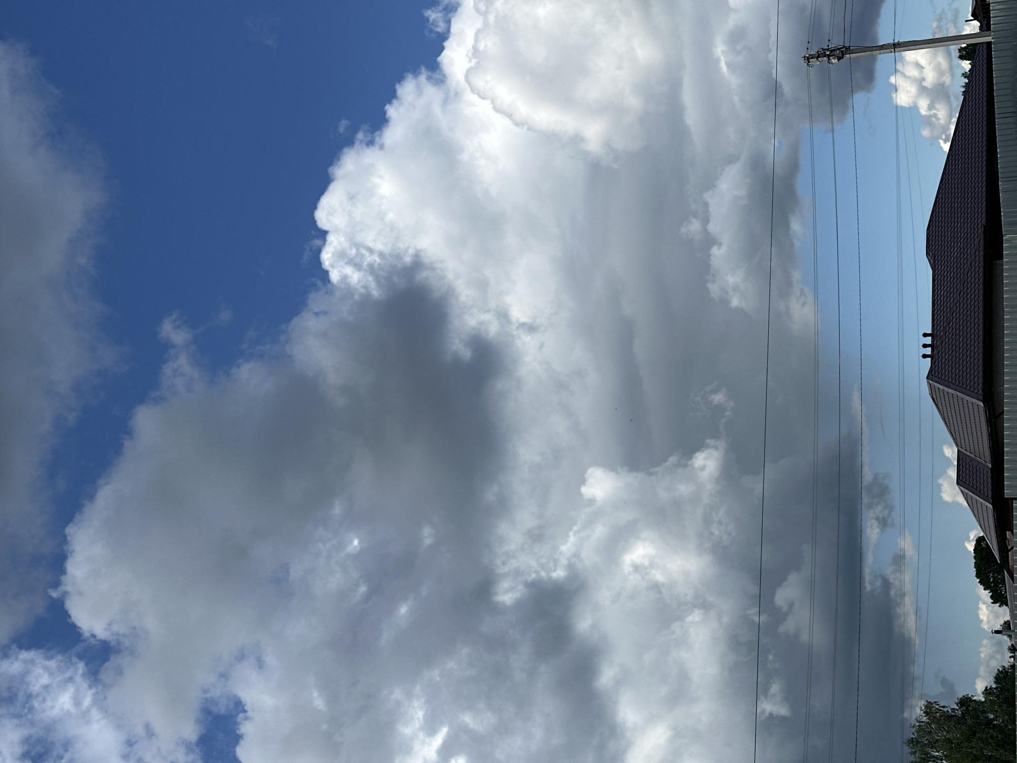 The image shows a view of the sky, with a mix of blue sky and large, billowing cumulus clouds. Some of the clouds are white and bright, while others are darker gray, suggesting varying densities and potential for precipitation. A power pole and several electrical lines are visible, running vertically across the frame. A portion of a building with a dark-colored roof is present on the right side of the image, with a few trees visible below the roof. The scene appears to be in Kharnavy, Russia, but no people or specific activities are visible in the image.