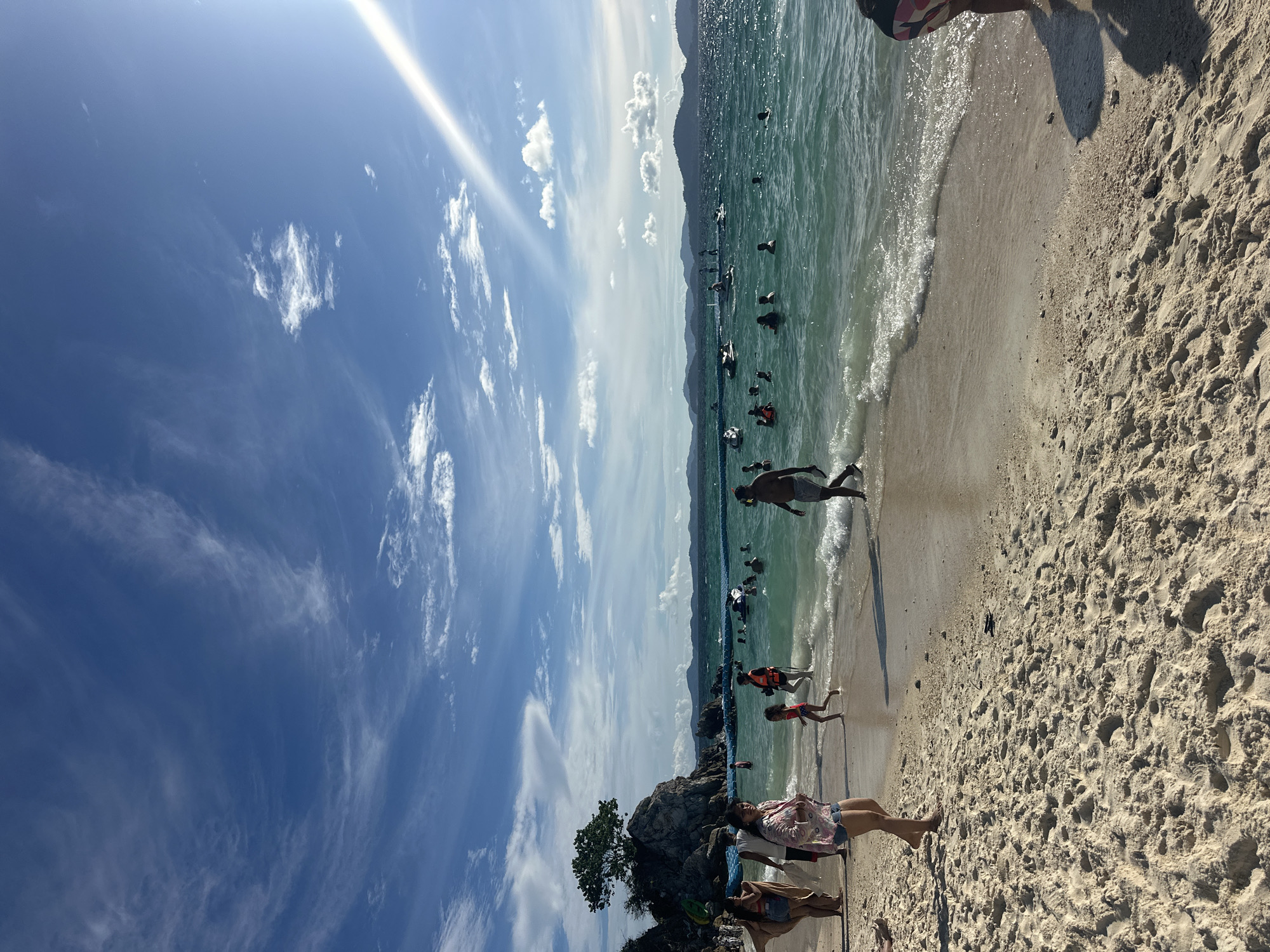 This image depicts a vibrant beach scene in Phru Nai, Thailand, likely during the day given the bright sunlight. The image is oriented vertically, showing a wide expanse of a sandy beach in the foreground, with gentle waves lapping at the shore. The water is a clear, turquoise green, with numerous people scattered throughout, some swimming, others floating or playing near the water's edge. Several small boats are visible further out in the sea.
In the background, a mountainous coastline can be seen, hinting at the tropical location. The sky is a brilliant blue, dotted with wispy white clouds, and a strong sunbeam cuts across the upper left of the frame, indicating a sunny and likely warm weather. A few individuals are walking along the beach in the foreground, with some appearing to be relaxing or posing. The overall mood is one of leisure and enjoyment, characteristic of a tropical vacation. There are no visible text elements in the image.