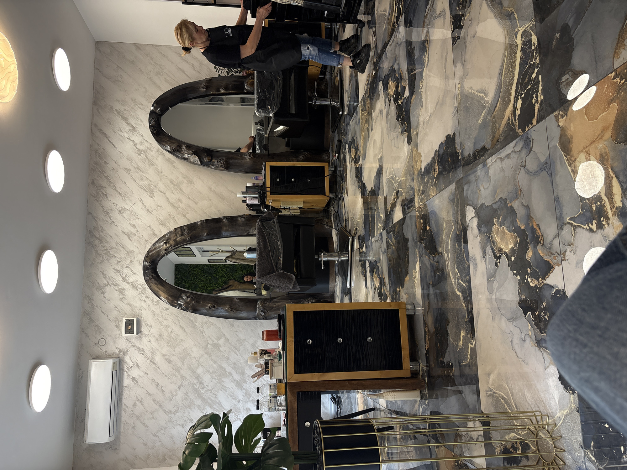 A brightly lit interior space, likely a hair salon, features two hairstyling stations. Each station includes an oval mirror framed with a dark, weathered material, a black styling chair, and a black and gold storage unit. Various styling products and bottles are visible on the storage units. The walls are decorated with a mottled gray and white pattern, and the floor is covered in large tiles with a swirling pattern of gray, black, and gold. One person, wearing a black top and jeans, is standing at one of the stations, seemingly working on a client whose reflection is visible in one of the mirrors. A large green plant is present in the foreground. The location is a business premise within Plovdiv, Bulgaria.