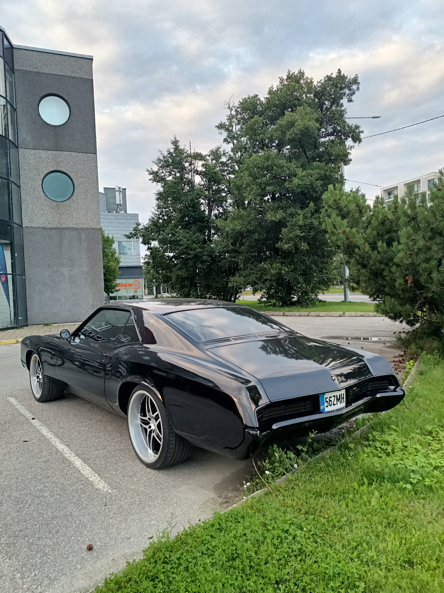 In Tallinn, Estonia, a sleek, black vintage car is parked on an asphalt lot. The car, likely a 1970s American coupe, features large, custom chrome wheels and a distinctive rear end with a license plate reading "56ZMH EST." It is positioned alongside a building with a textured grey facade and large, circular windows, suggesting an office or commercial structure. Lush green grass and trees surround the parking area, with a street and more buildings visible in the background under an overcast sky. The scene appears to be a typical urban environment, possibly a business district or a quiet street. There are no people visible in the image.