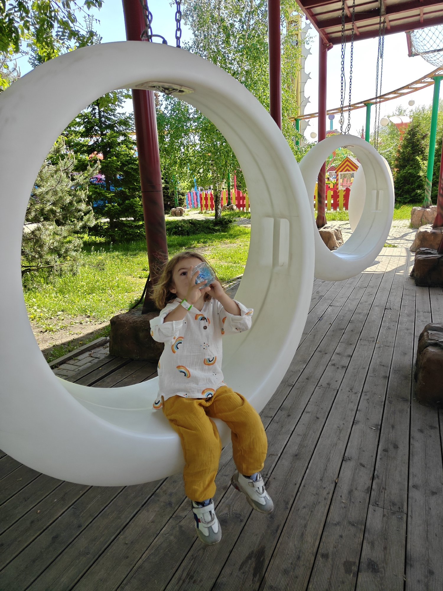 A young child is drinking from a blue bottle while sitting on a large, white circular swing. The swing is located on a wooden deck at a park or playground, identified as Khimki, Russia, based on the availability of location information. The child is wearing a white shirt with rainbow designs and mustard-colored pants, along with athletic shoes. The scene is outdoors and includes additional swings, trees, and playground elements like colorful fences. The child has a wristband. The area around the swing is green with grass and some decorative landscaping.