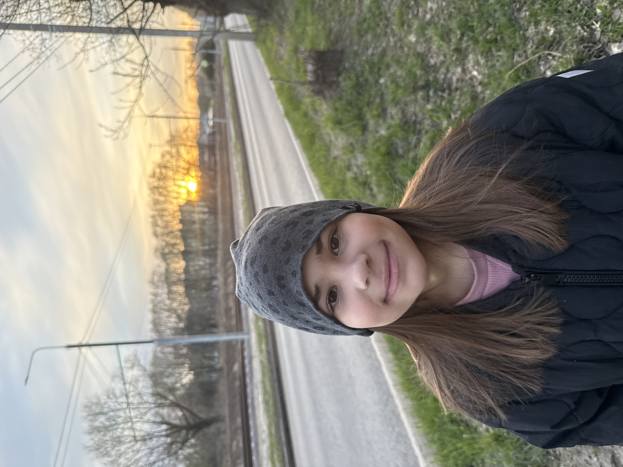 A young girl in a dark jacket and spotted grey hat smiles at the camera. She is standing on the edge of a road in Rusanovo, Russia, next to a grassy area. Behind her, across the road, are tram tracks and a row of trees with a sunset visible through the branches.