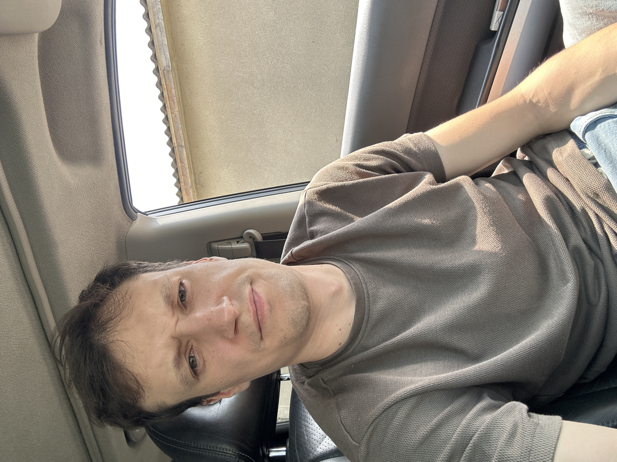 A young man with brown hair is sitting in the passenger seat of a car, looking at the camera with a slightly furrowed brow. He is wearing a dark olive-green short-sleeved shirt and blue jeans. The interior of the car is visible, including a black leather headrest and a gray headliner. Sunlight streams in through the window, casting shadows on the man's face and arm. Outside the car window, a beige wall and a corrugated roof are visible. The overall scene appears to be outdoors in a suburban or rural setting. The city of Shatava, Ukraine is not depicted or suggested by any visual cues in the image.
