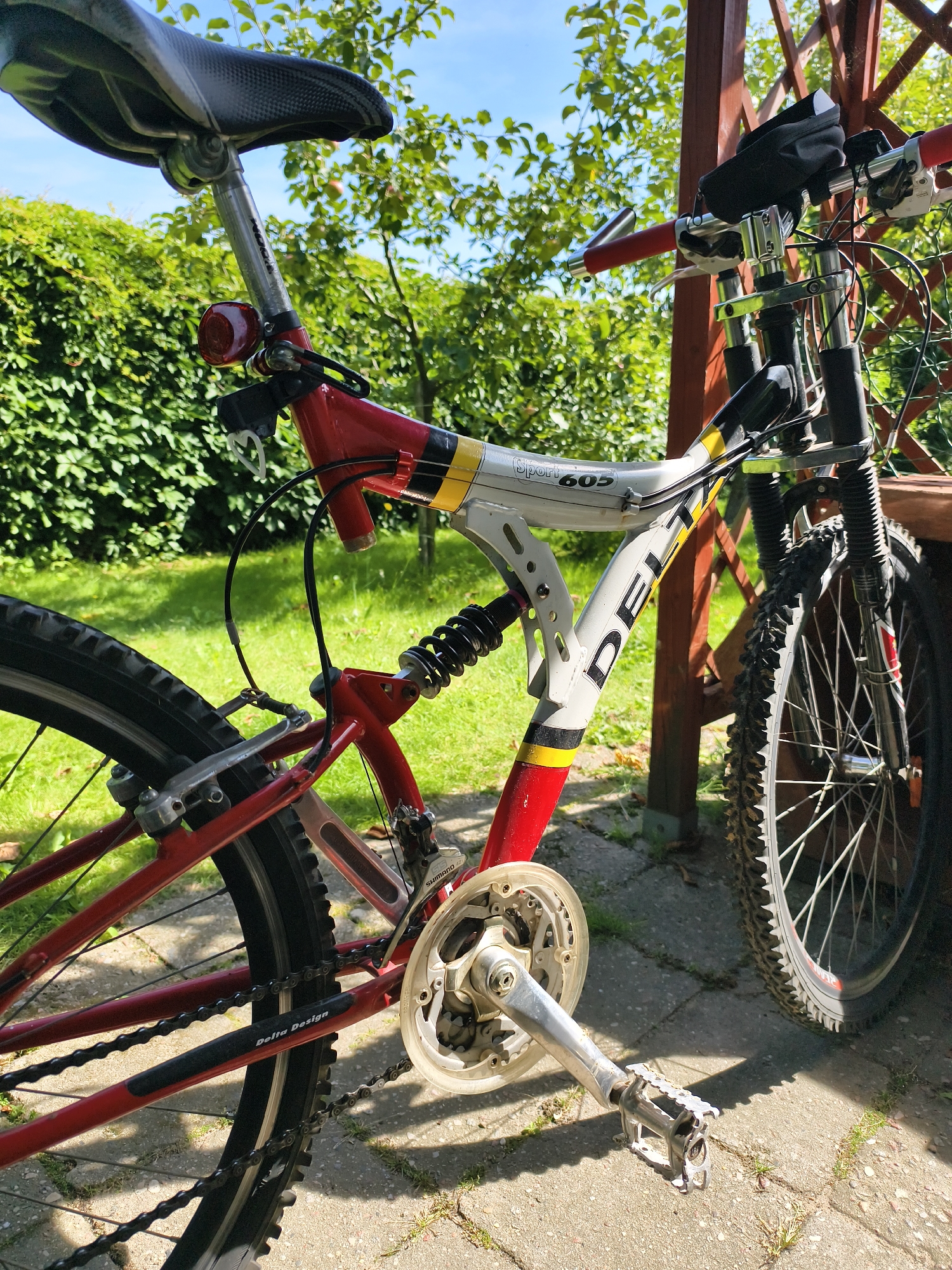 The image shows a red and white full-suspension bicycle parked outdoors on a paved surface with grass and vegetation visible in the background. The bicycle is angled towards the viewer's right, with its rear wheel and part of the frame prominently displayed. The seat is black with a carbon fiber pattern. A red taillight is attached to the seat post. The frame has "DELTA" and "Sport 605" text. The bicycle has a front suspension fork, handlebars with red grips, and a small black bag attached to the handlebars. The front tire is knobby. The rear wheel has a derailleur and chain. The overall setting suggests a sunny day in a residential or recreational area. The image is not specifically located in Tczew, Poland.