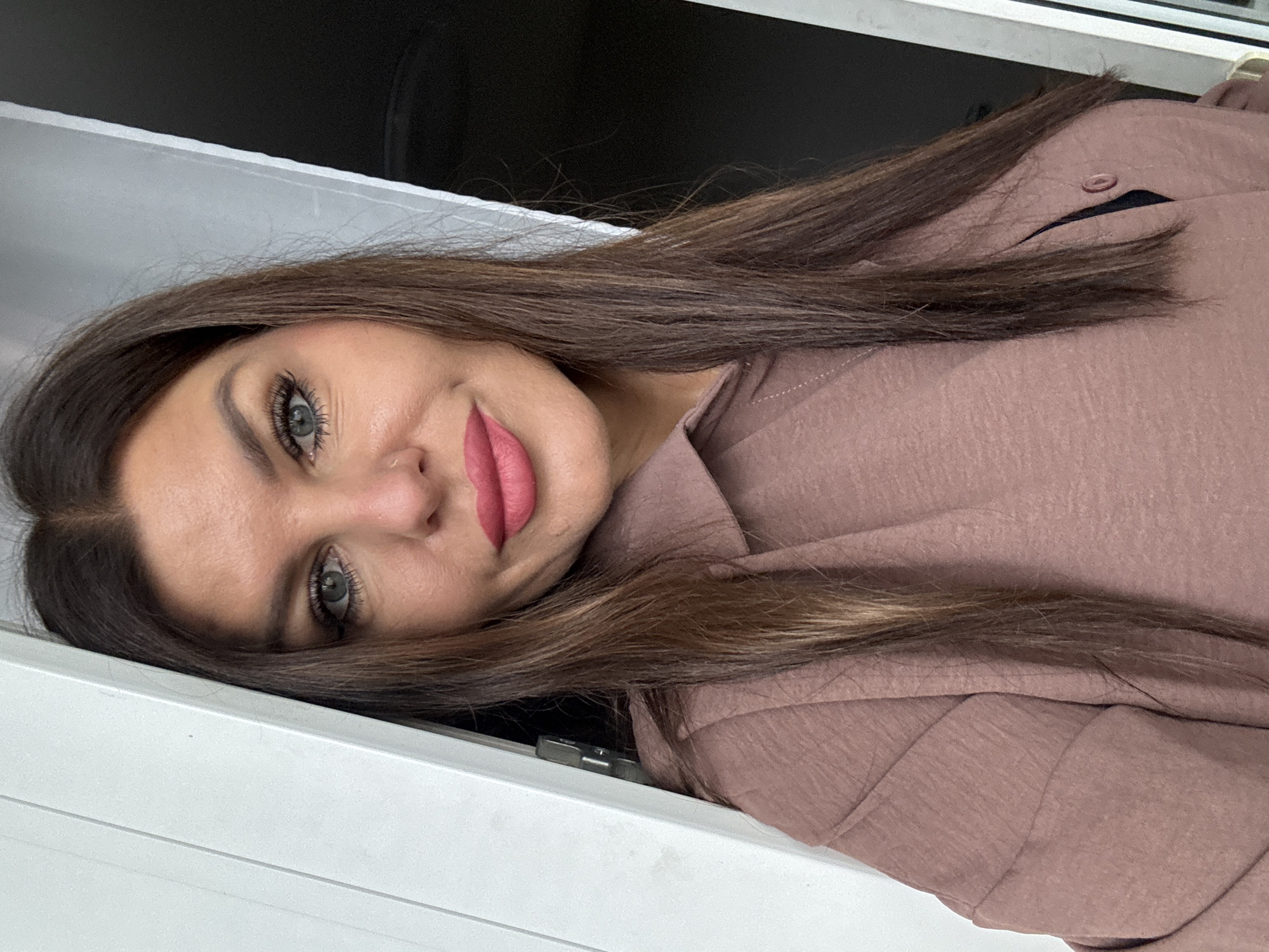 A woman with long, brown hair and striking blue eyes is looking directly at the camera. She is wearing a mauve-colored collared shirt. Her lips are painted a vibrant pink. The woman is positioned in a doorway, with a white door frame to her left and a dark background to her right. The scene does not appear to be in Chaikovsky, Russia.