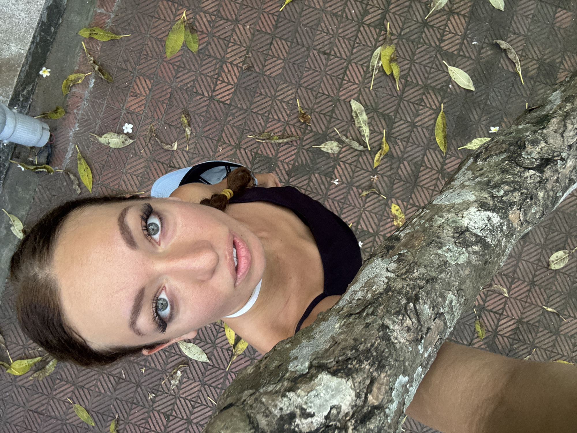 The image depicts a woman looking up at the camera from a low angle. She has fair skin, brown hair in a ponytail with a yellow band, and blue-grey eyes. She is wearing a black top and a white necklace. Part of a tree trunk obscures a portion of the lower right of the frame. The background is a brick-patterned ground covered with fallen leaves and small white flowers. A gray container is partially visible in the upper left. The location appears to be in Hà Nội, Vietnam.