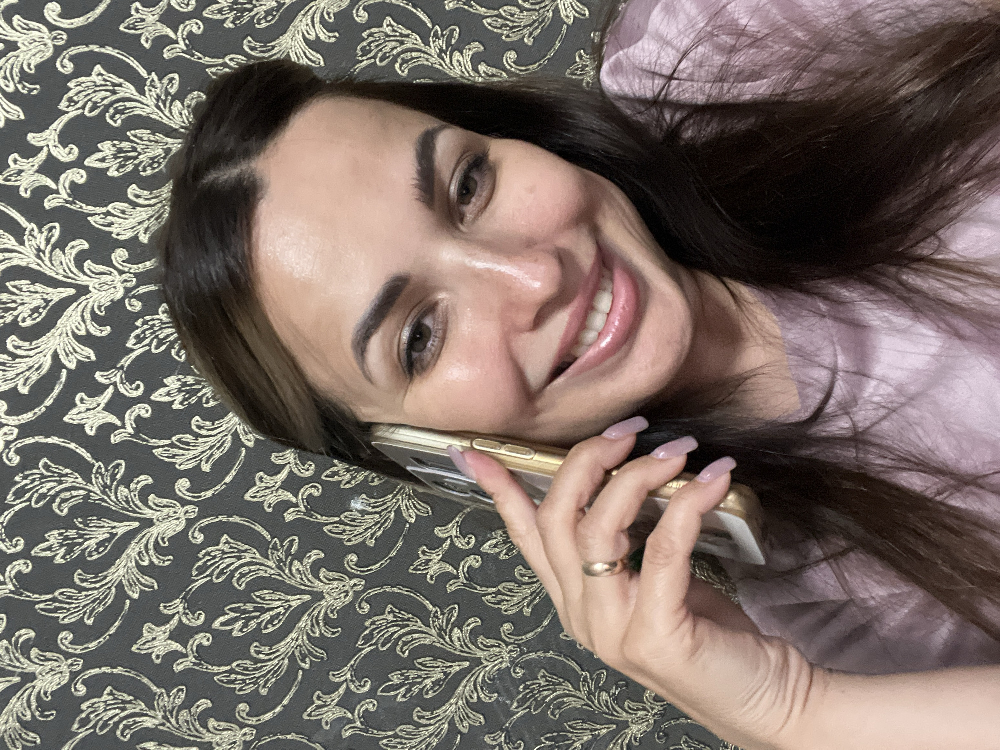 A woman with dark brown hair, wearing a light purple shirt and a gold ring on her left hand, is holding a gold cell phone to her left ear. She is smiling and looking at the camera. The background is a dark gray wallpaper with a repeating gold floral pattern. The setting is indoors.