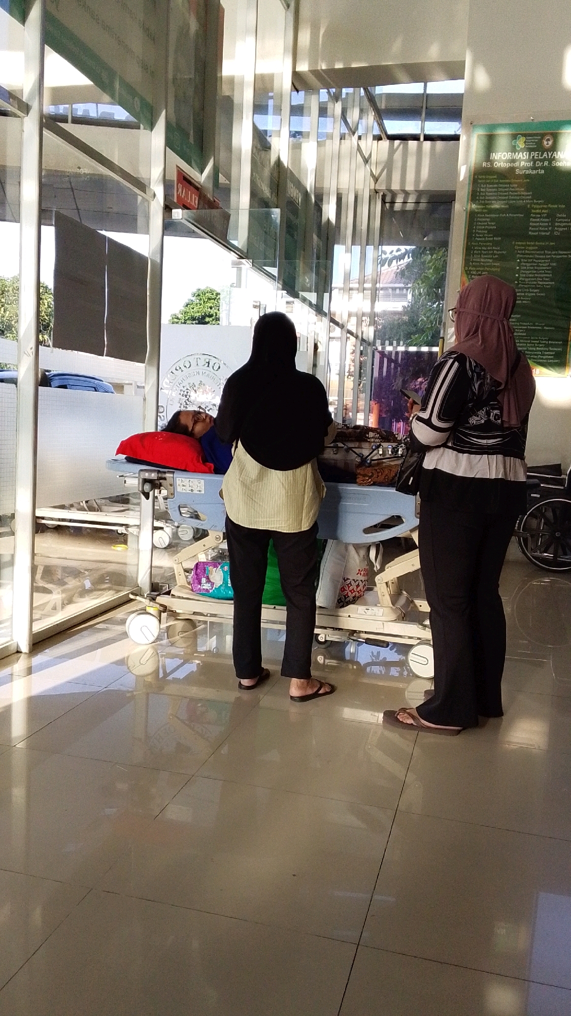 In Pabelan, Indonesia, inside a hospital lobby, a patient lies on a stretcher. Two women stand nearby, one in a black hijab and a beige tunic, the other in a mauve hijab and a black and white patterned jacket. The patient is lying on their back on a blue and white hospital bed, propped up by a red pillow. The floor is polished beige tile, reflecting the bright, natural light filtering through large glass windows and doors. A large green informational poster is visible on the right wall. The overall scene suggests a waiting area or a pathway within the hospital.