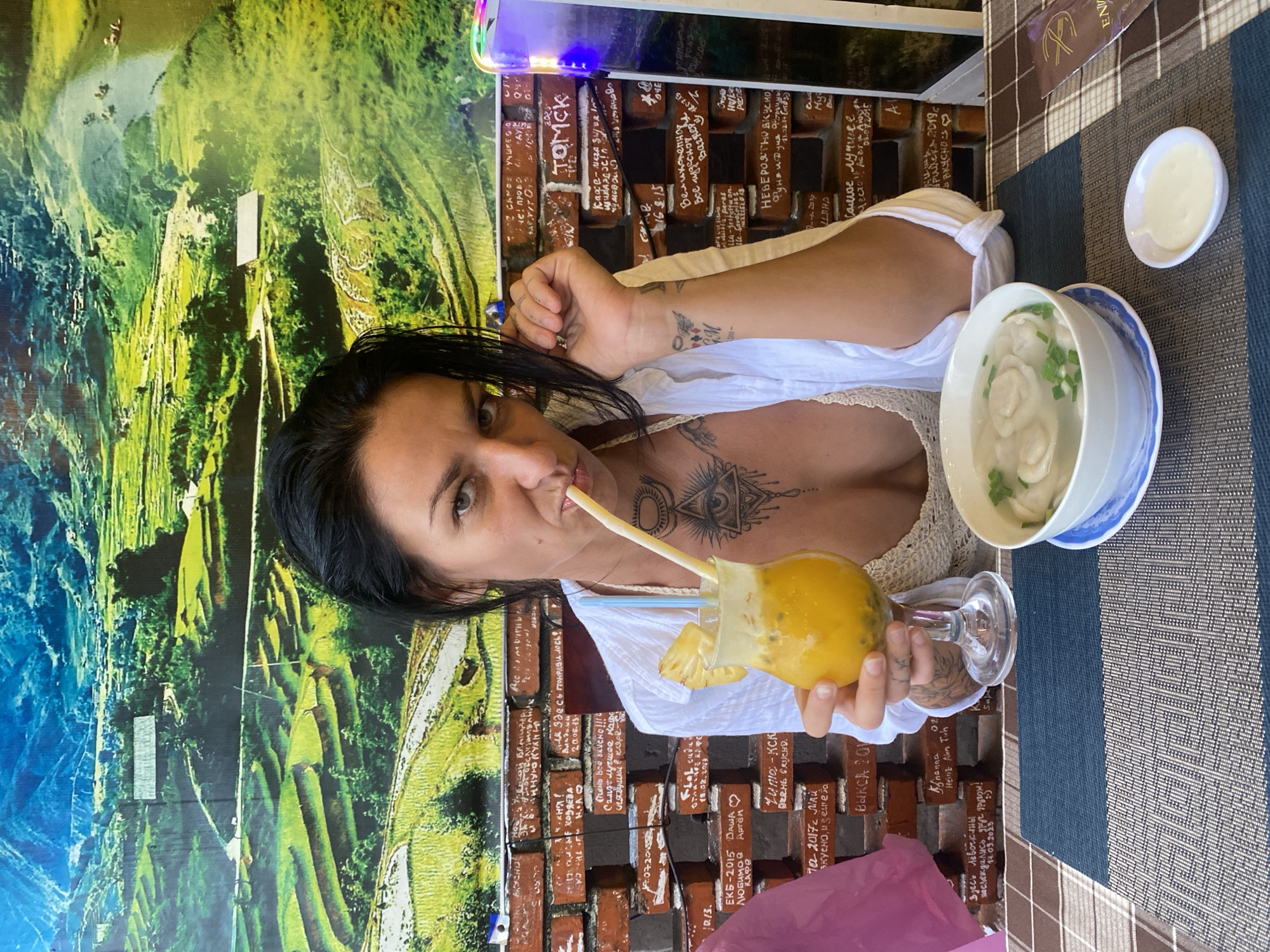 The image shows a woman at a table in what appears to be a restaurant in Phu Thuy Ward, Vietnam. She is fair-skinned, with dark hair, and is drinking a yellow beverage with a straw. She has tattoos on her chest, neck, and arm. On the table, there is a bowl of food that looks like soup with dumplings, a small dish with a white sauce, a pink bag, and what appears to be a menu. The table has a checkered tablecloth with blue and tan stripes. The walls behind the woman are decorated with a mural of green rice terraces and a brick-like pattern with writing in what seems to be Russian.