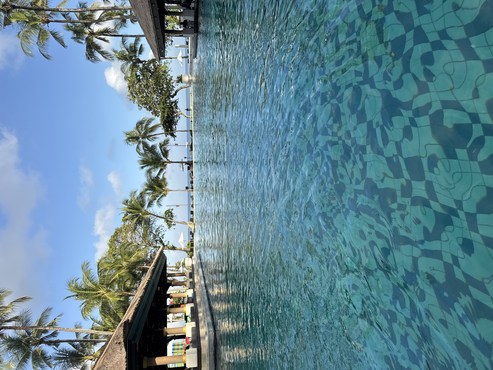The image shows a swimming pool next to a structure with a thatched roof, likely a beachside resort in Jimbaran, Indonesia. Palm trees line the edge of the pool. The water in the pool is turquoise and the bottom has a checkered pattern. The sky is blue with some clouds.