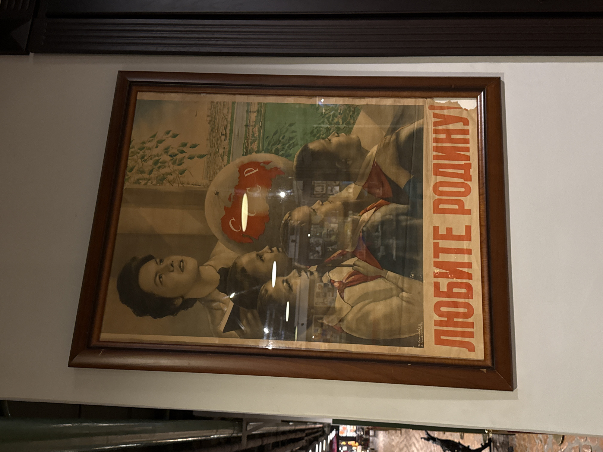 The image shows a framed Soviet-era propaganda poster hanging on a white wall. The poster depicts a group of young people, likely Pioneers, looking towards a globe and a cityscape. The globe is red and prominently displays the letters "СССР" (USSR). The cityscape appears to be a stylized representation of a Russian city, possibly Moscow, with a river and buildings. The overall theme of the poster is patriotism and love for the homeland. The phrase "ЛЮБИТЕ РОДИНУ!" (Love the Motherland!) is printed in large red letters at the bottom of the poster. The poster is slightly worn, with some damage to the paper. The framing is simple and made of dark wood. The background of the image suggests an indoor setting, possibly a museum or a public building in Arkhangel'skoe, Russia, given the context of Soviet-era art.