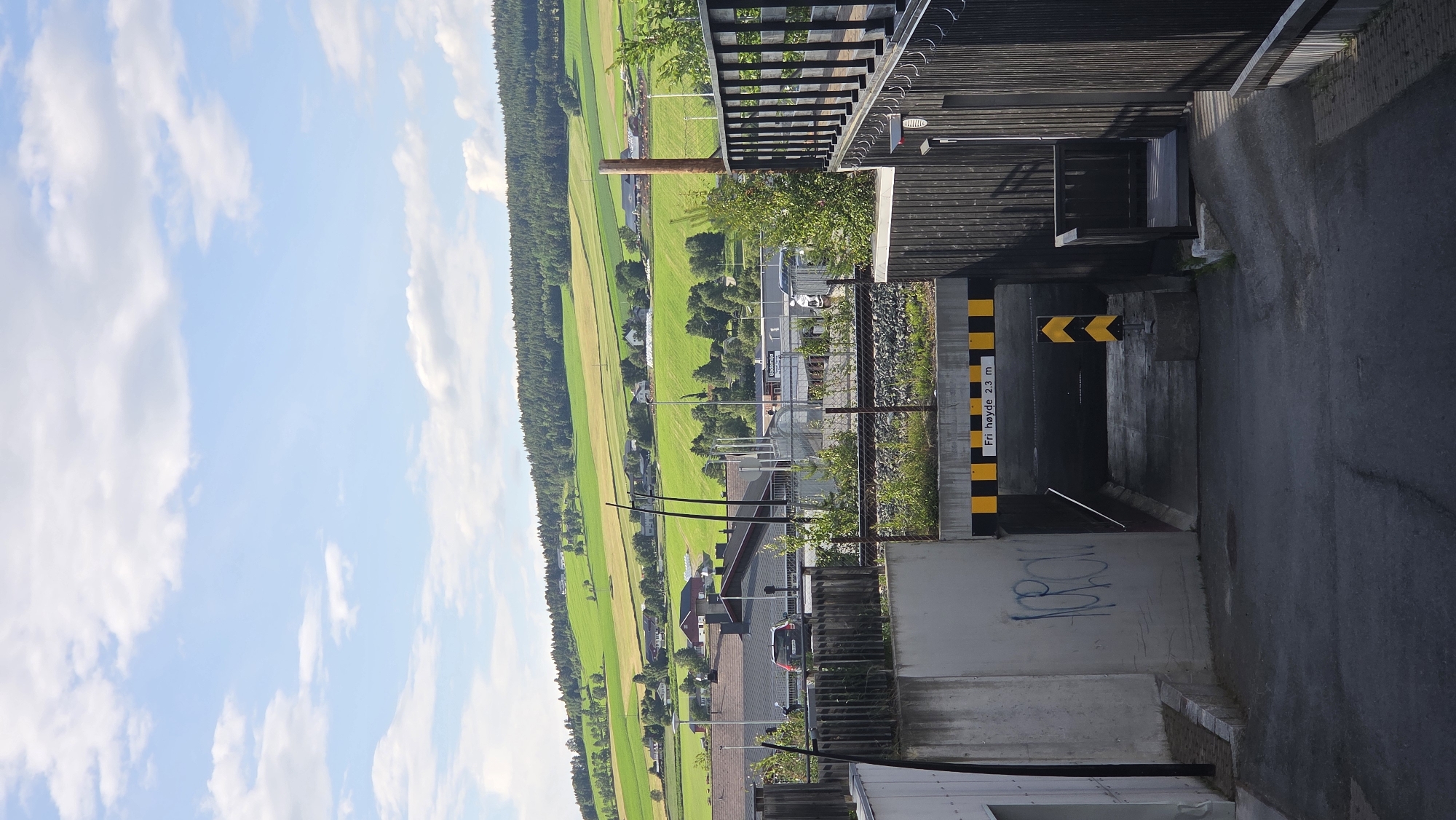 The image shows a downward sloping road leading to an underpass, marked with a sign indicating a free height of 2.3 meters. The underpass has yellow and black striped barriers and a yellow chevron sign. To the left of the underpass is a concrete wall with graffiti. To the right is a dark wooden fence with railings. In the background, there are train tracks, buildings, and a scenic landscape of rolling green hills with scattered houses under a blue sky with white clouds. The scene is likely located in Tynset, Norway, given the Norwegian text on the height restriction sign. There are no people visible in the image.