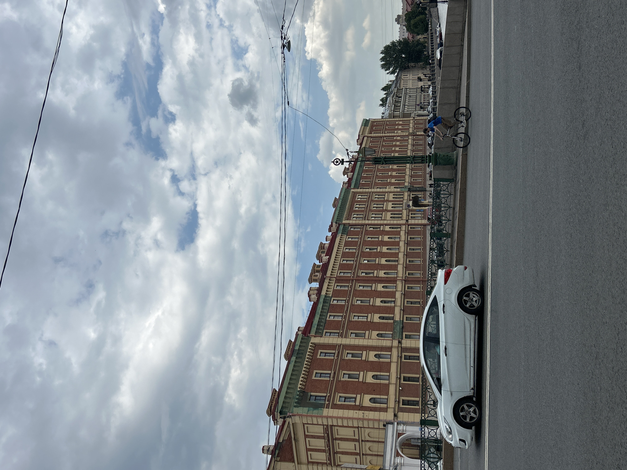 A white sedan drives along a paved road in front of a multi-story brick building with a red roof. The building features numerous windows and decorative elements. A cyclist wearing a blue shirt and a backpack rides on the sidewalk parallel to the road. Overhead wires are visible against a cloudy sky. The scene appears to be in an urban environment with a canal or river in the background.