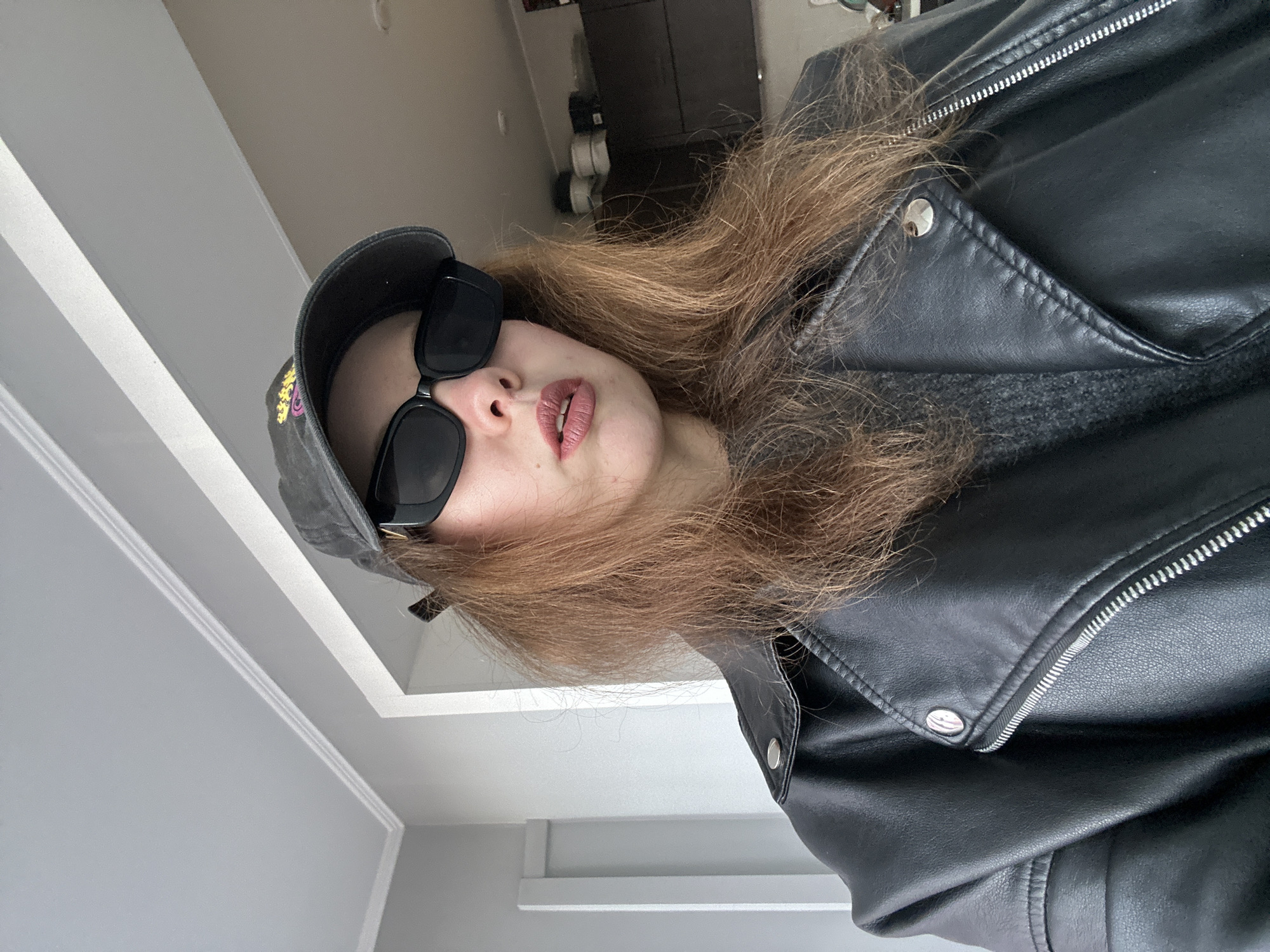 The image is a close-up selfie of a person wearing a black baseball cap, black sunglasses, and a black leather jacket. The person has medium-length brown hair and visible lips. The background is a blurred interior with a white wall and what appears to be a part of a ceiling. There is no visible indication that the location is in the city of Krasnoyarsk, Russia.