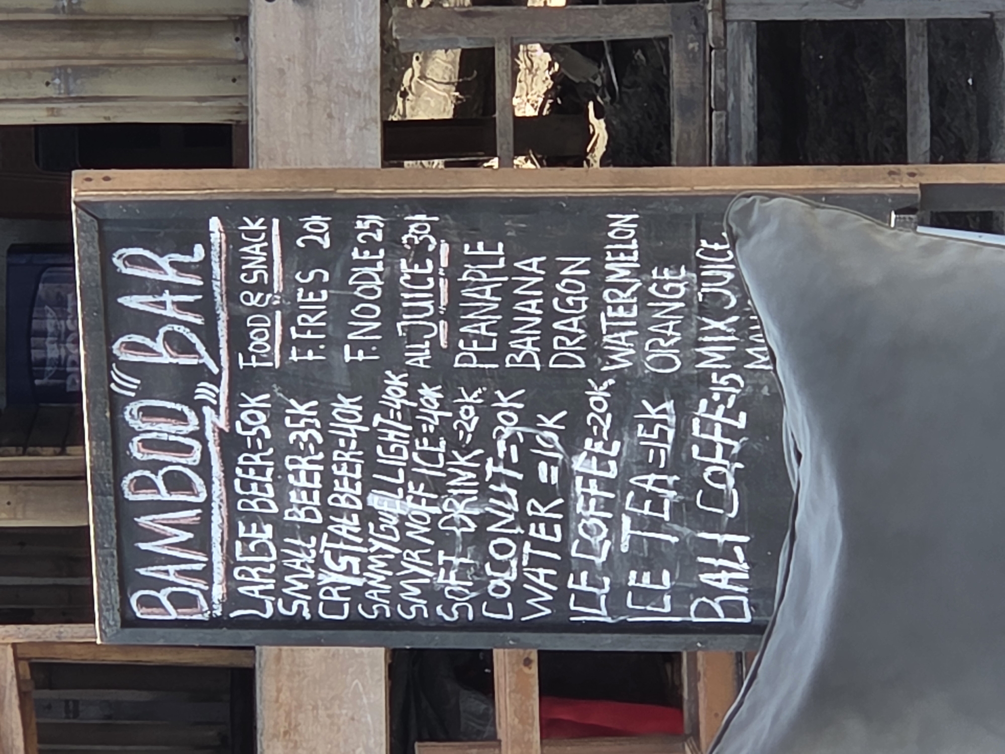 The image shows a chalkboard sign for "Bamboo Bar," listing prices for various food and drink items. The sign appears to be mounted on a wooden frame, with a partially visible structure behind it. The listed items include "Large Beer - 50K," "Small Beer - 35K," "Crystal Beer - 40K," "Sammyguel Light - 40K," "Smyr Noffice - 40K," "Soft Drink - 20K," "Coconut - 30K," "Water - 10K," "Ice Coffee - 20K," "Ice Tea - 15K," and "Bali Coffee - 15," along with food options like "F. Fries 20," and "F. Noodle 25." The sign also lists "All Juice - 30," and various fruit options like "Peanaple," "Banana," "Dragon," "Watermelon," "Orange" and "Mix Juice" The sign is likely from North Kuta, Indonesia.