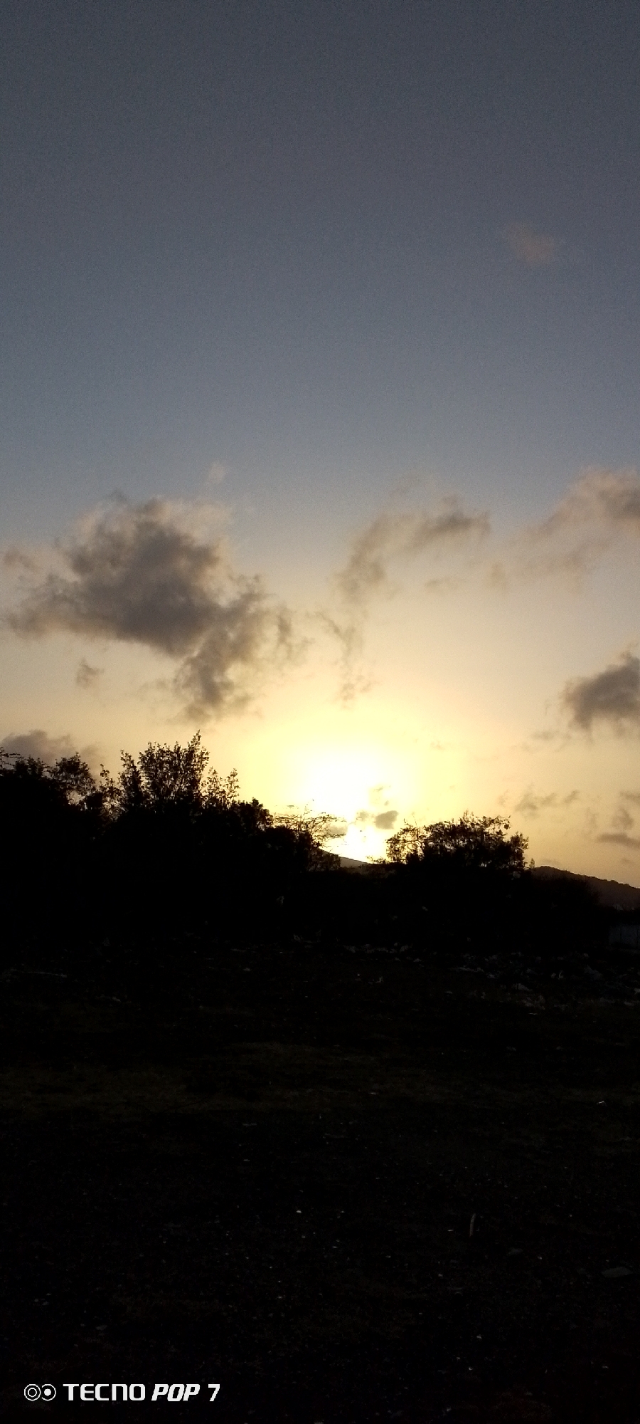 The image shows a sunset in Altagracia, Venezuela. The sky is a gradient of blue to yellow, with dark clouds obscuring the setting sun. Below, there is a dark silhouette of trees, and the ground is mostly obscured by darkness. "TECNO POP 7" is visible in the lower-left corner of the image.