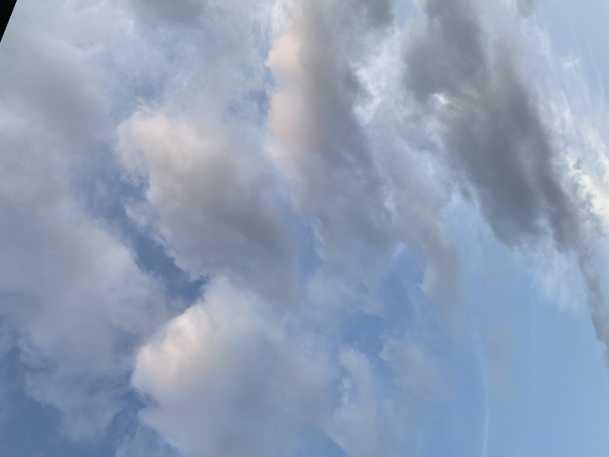 A wide shot of a sky filled with clouds is presented. The clouds are varied in color, ranging from bright white to light pink and dark gray. The sky is a soft blue, with some areas showing lighter patches where the clouds are thinner. The lighting suggests it might be late afternoon or early evening. No specific city or landmarks are visible, only the sky and clouds.