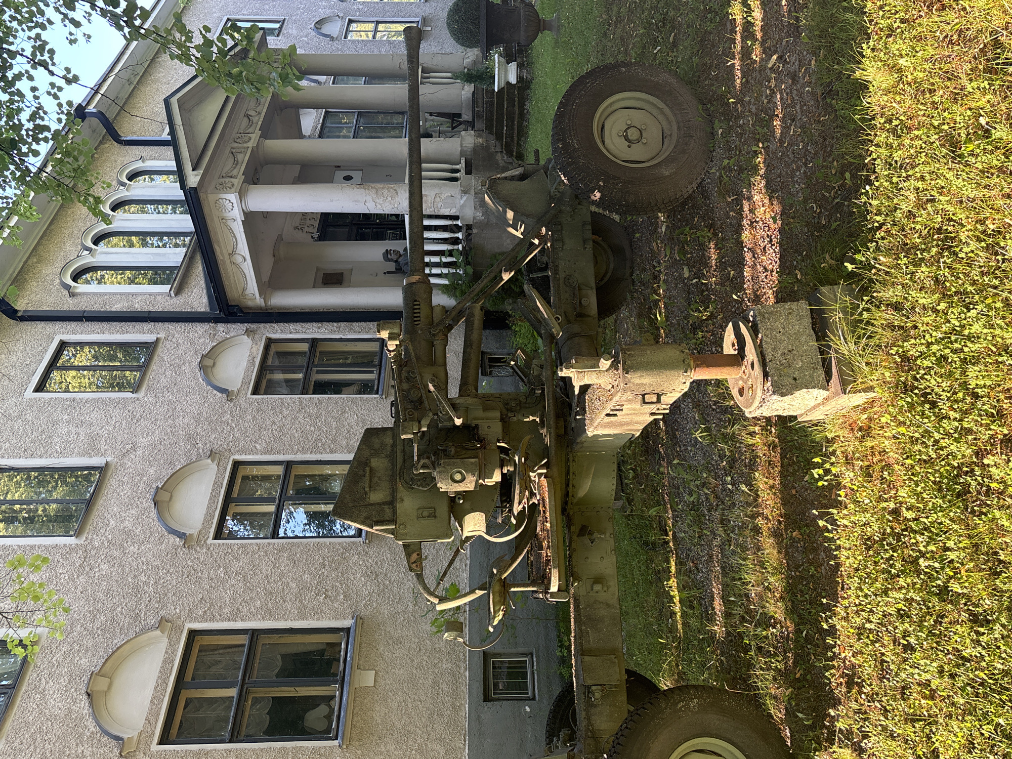 The image depicts a disused anti-aircraft gun mounted on a towed carriage, situated in front of a building. The gun is green and appears weathered, with visible rust and wear. It is positioned on a patch of grass and gravel, with shadows from overhead trees indicating it is daytime. The building behind the gun is a two-story structure with a light-colored, textured facade. It features several windows, some with arched tops, and a prominent portico with columns. The overall impression is one of historical preservation or display, given the placement of the military artifact in front of a significant building. The location is the Kruununpuisto Park in Imatra, Finland, specifically near the Imatrankoski rapids.