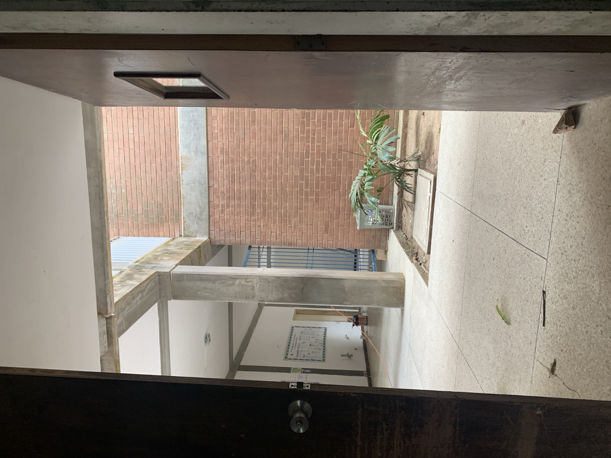 The image shows a view through a doorway in Las Piedras, Venezuela, leading to an outdoor courtyard. A red brick wall is visible on the left, with a concrete overhang and supporting columns beneath. A plant sits in a container near a drain in the courtyard. The flooring is light-colored, tiled concrete. A blue metal gate is visible further into the courtyard. Inside the building, a whiteboard is affixed to the wall. A door handle is in the foreground, indicating a partially open door.