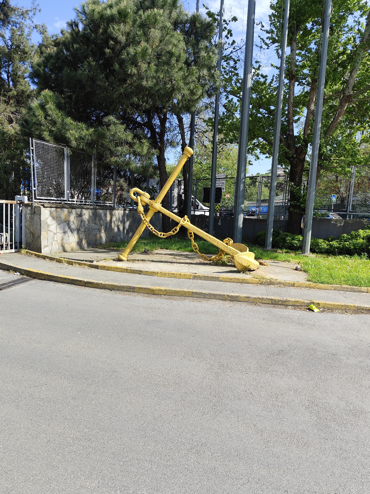 The image shows a large, yellow-painted anchor with a chain, displayed on a concrete platform at the edge of a road. Behind the anchor, there is a low wall made of stone and metal fencing, with trees and greenery in the background. Several tall, grey metal poles are also visible, possibly flagpoles. The location appears to be an urban or suburban area with well-maintained landscaping.