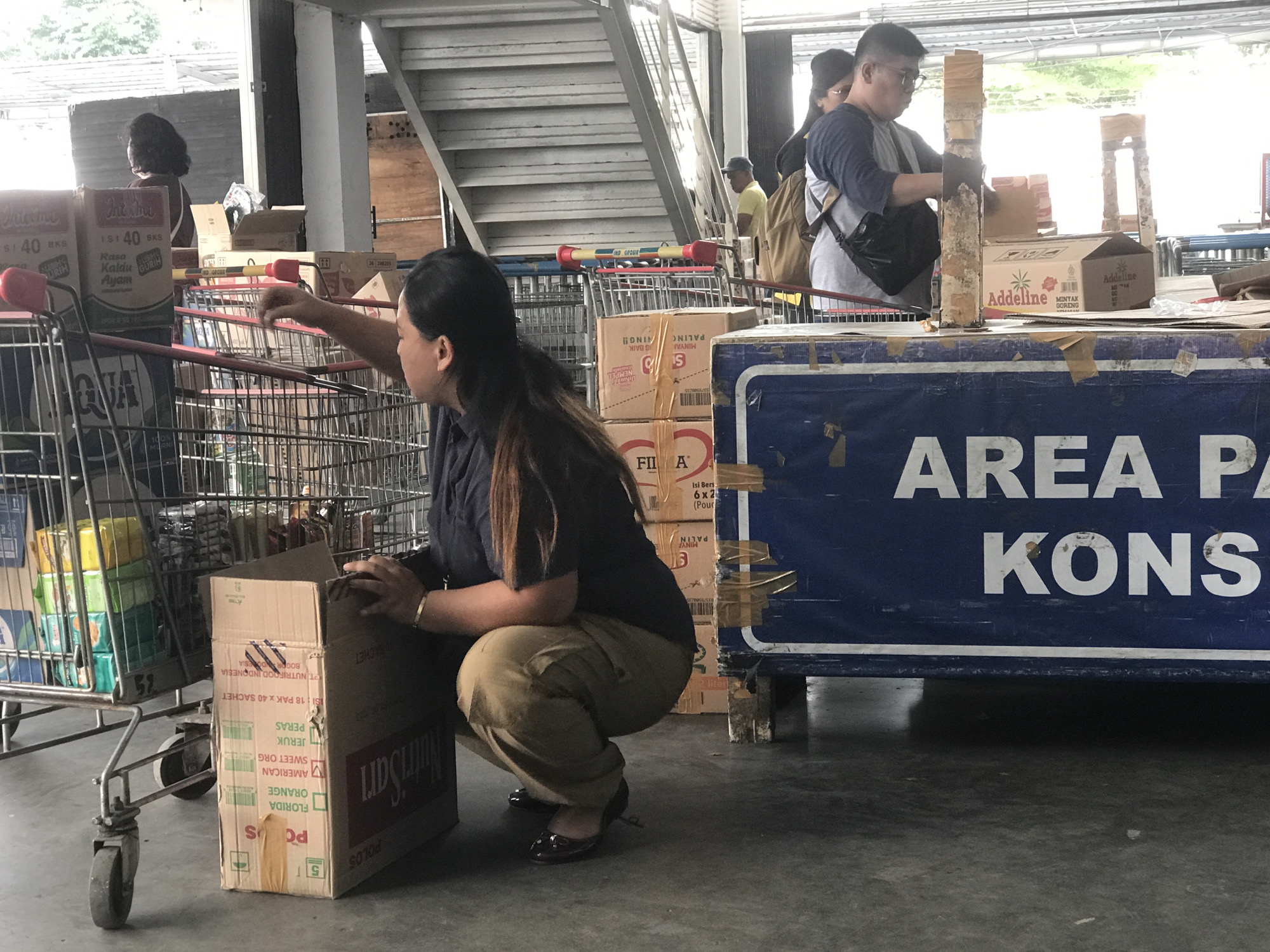 Outside what appears to be a wholesale grocery store, a woman with dark hair is crouched down next to a shopping cart, placing items from a cardboard box into it. The cart already contains various food and beverage products. A second shopping cart sits to the left of the cart the woman is loading. Stacks of boxed goods are behind the woman and next to the second shopping cart. In the background, other shoppers and the entrance of the store can be seen. A blue sign reading "AREA PA KONS" is in the bottom right of the image. Text on boxes identifies the location as Bogor, Indonesia. Other text on boxes in the image show Indofood and Nutrisari branding.