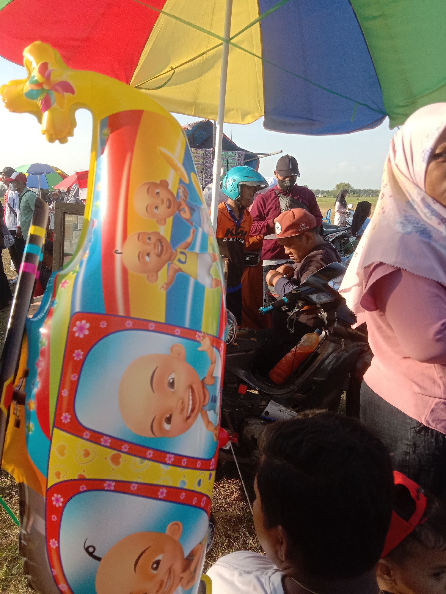 A group of people are gathered outdoors, possibly at a local event or market, with several large colorful umbrellas providing shade. In the foreground, a brightly colored inflatable object, possibly a toy, with cartoon faces is prominently displayed. Behind it, several individuals are visible. One person wears an orange shirt and a teal helmet, while another, wearing a maroon shirt and a black cap, appears to be interacting with a motorbike. A woman in a pink hijab is also present. The environment seems to be an open field, suggesting a rural or semi-urban setting. There are glimpses of other people and stalls in the background, indicating a public gathering. The lighting suggests it is daytime, with the sun casting shadows.