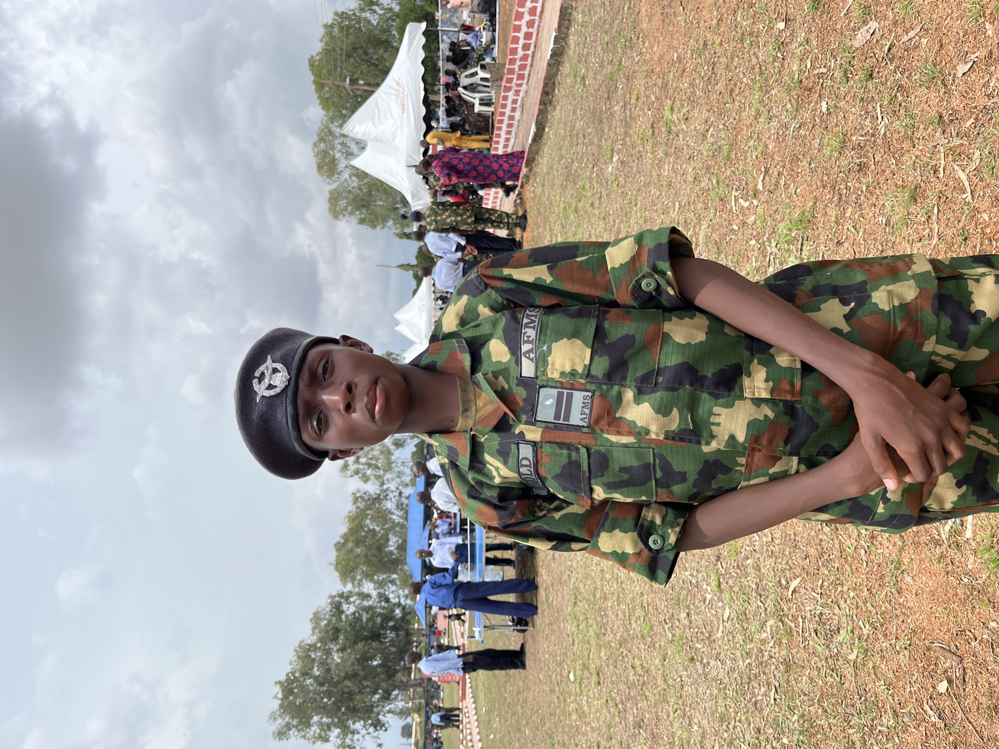A young person wearing a camouflage uniform and a black beret stands in the foreground, facing the camera. They have their hands clasped in front of them. The uniform has patches on the sleeves; one reads "OLD" and the other "AFMS". In the background, there are white tents, people in civilian clothing and uniforms, and a ping pong table. The setting appears to be an outdoor event or gathering, possibly a military ceremony or sporting event, with trees and a cloudy sky visible. The ground is covered in dry grass and dirt. The location is identified as Barakin Lerawi, Nigeria.