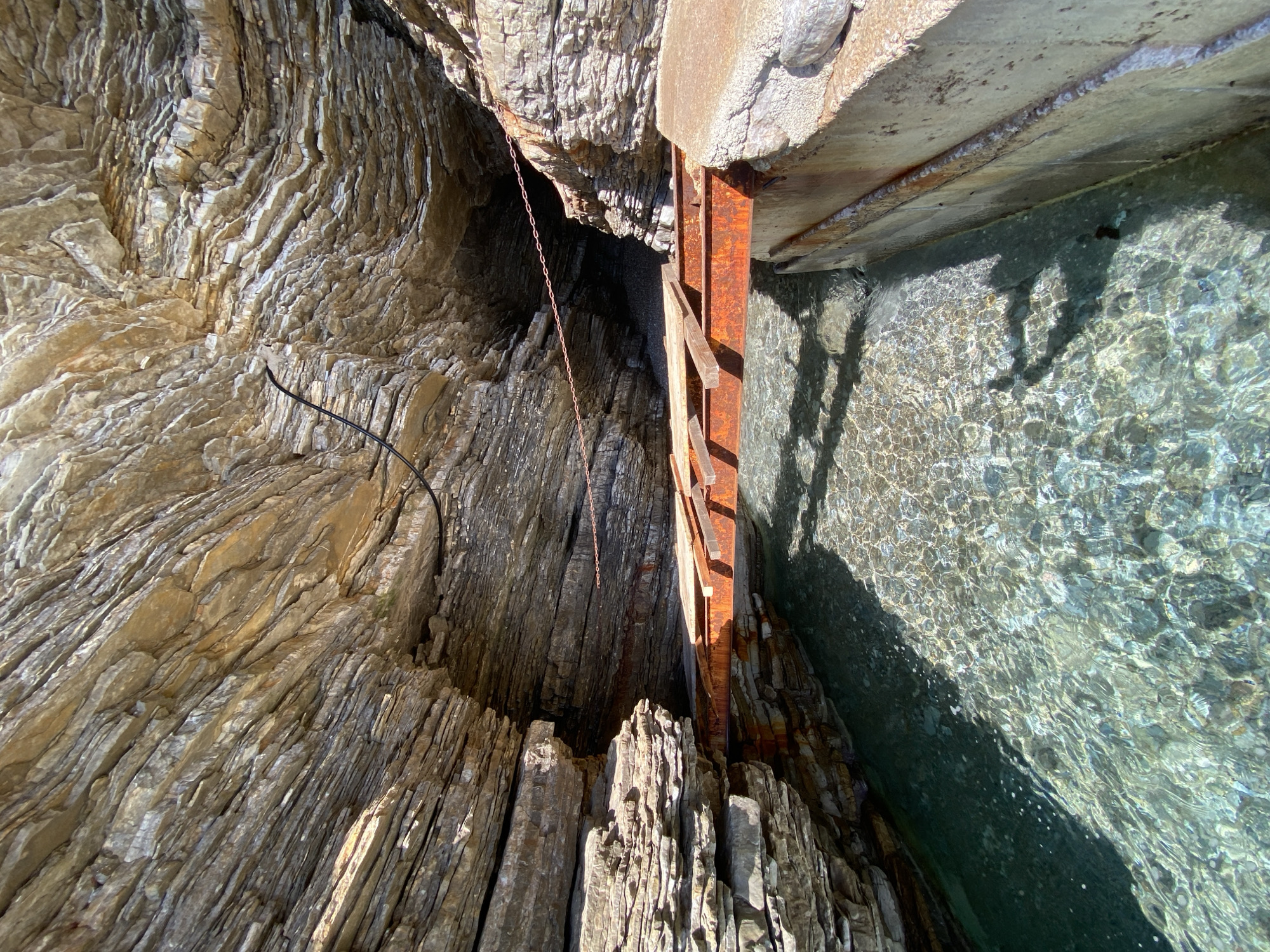 The image captures a geological formation and man-made structure in Sveti Stefan, Montenegro. A layered rock cliff face of sedimentary rock forms the left side of the frame. A rusty metal ladder descends vertically between the rock face and a concrete structure. The ladder appears old and weathered. Water fills the space on the right side of the frame. A hanging chain is attached to the rock face above the ladder. The water appears clear. A dark shadow is visible on the water's surface. A wire is partially visible on the rock face.