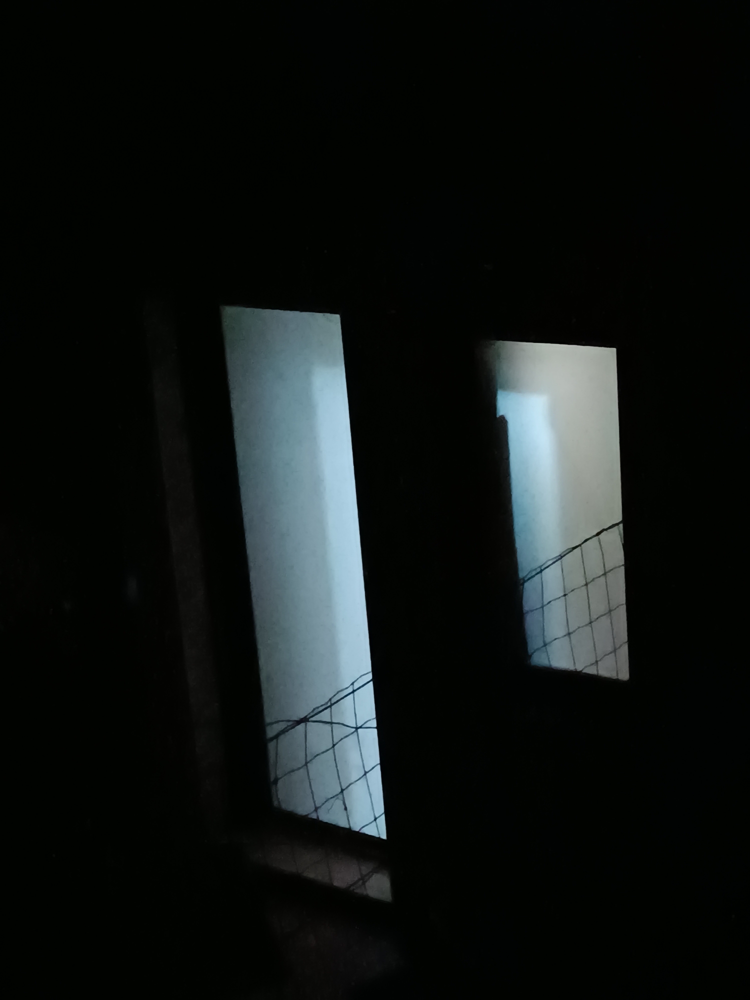 The image depicts a stark contrast between a dominant, deep black foreground and two illuminated rectangular openings. These openings, possibly windows or doorways, are seen from a very dark interior, suggesting night or an unlit room. The left opening is larger and appears closer, while the one on the right is smaller and set further back.
Through both openings, a bright, cool blue-white light is visible, illuminating what appears to be a light-colored surface or wall beyond. A distinctive dark, diamond-patterned net or grating stretches across the lower portion of these illuminated areas. This netting suggests a barrier, such as a fence or a sports net. The angle of the openings and the net creates a dynamic perspective against the overwhelming darkness. No people or specific activities are visible, and there is no discernible text. The scene is set in Viadana, Italy.