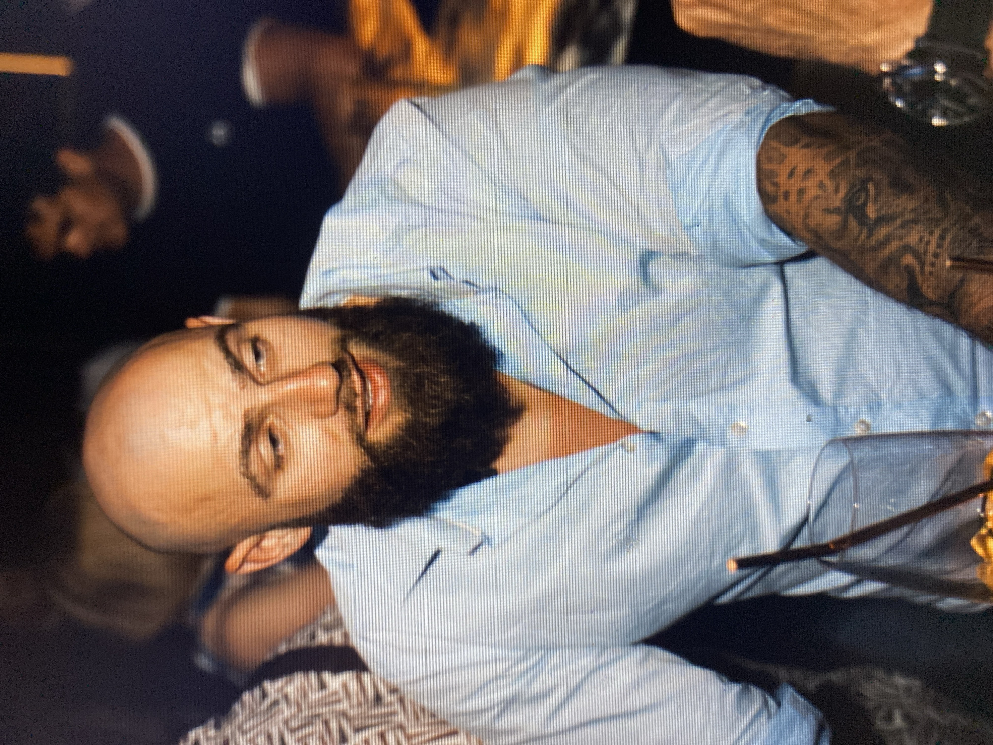 The image captures a bald man with a full, dark beard and light-colored eyes, prominently featured at a significant tilt. He is wearing a light blue, collared, button-up shirt. His right arm, visible on the left side of the frame, is covered in extensive, dark, intricate tattoos that are not clearly legible. The man is looking directly towards the viewer with a slight smile or smirk, suggesting a relaxed or positive mood.
In the background and foreground, blurred figures are visible, indicating a social gathering. A partial figure on the far left appears to be wearing a dark suit jacket with a white cuff. Another person is partially seen at the bottom, wearing a patterned garment. To the man's right, a clear glass containing a dark straw and yellowish liquid is visible.
The setting appears to be an indoor or evening social event, such as a bar, restaurant, or party, characterized by dim, artificial lighting and blurred, warm lights in the upper background. No specific visual cues within the image confirm the provided location context of Bankya, Bulgaria. The image shows noticeable pixelation. No discernible text is visible on any subject or object, including the tattoos.