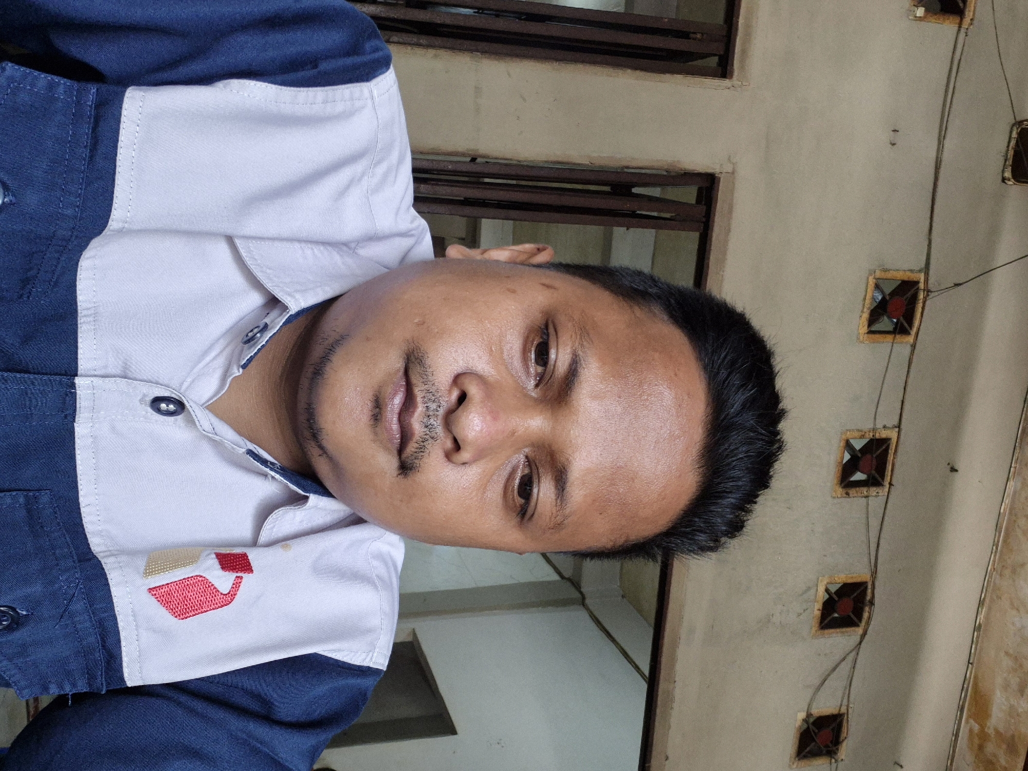 A medium shot depicts a man in a blue and white shirt, potentially indoors. The background shows a wall with several square decorative elements that have a red dot in the middle. Above the man, there appears to be a barred window or similar structure. The overall scene does not provide enough information to definitively place the image in Lhokseumawe, Indonesia. There are no clear landmarks or environmental characteristics that would strongly suggest that location.