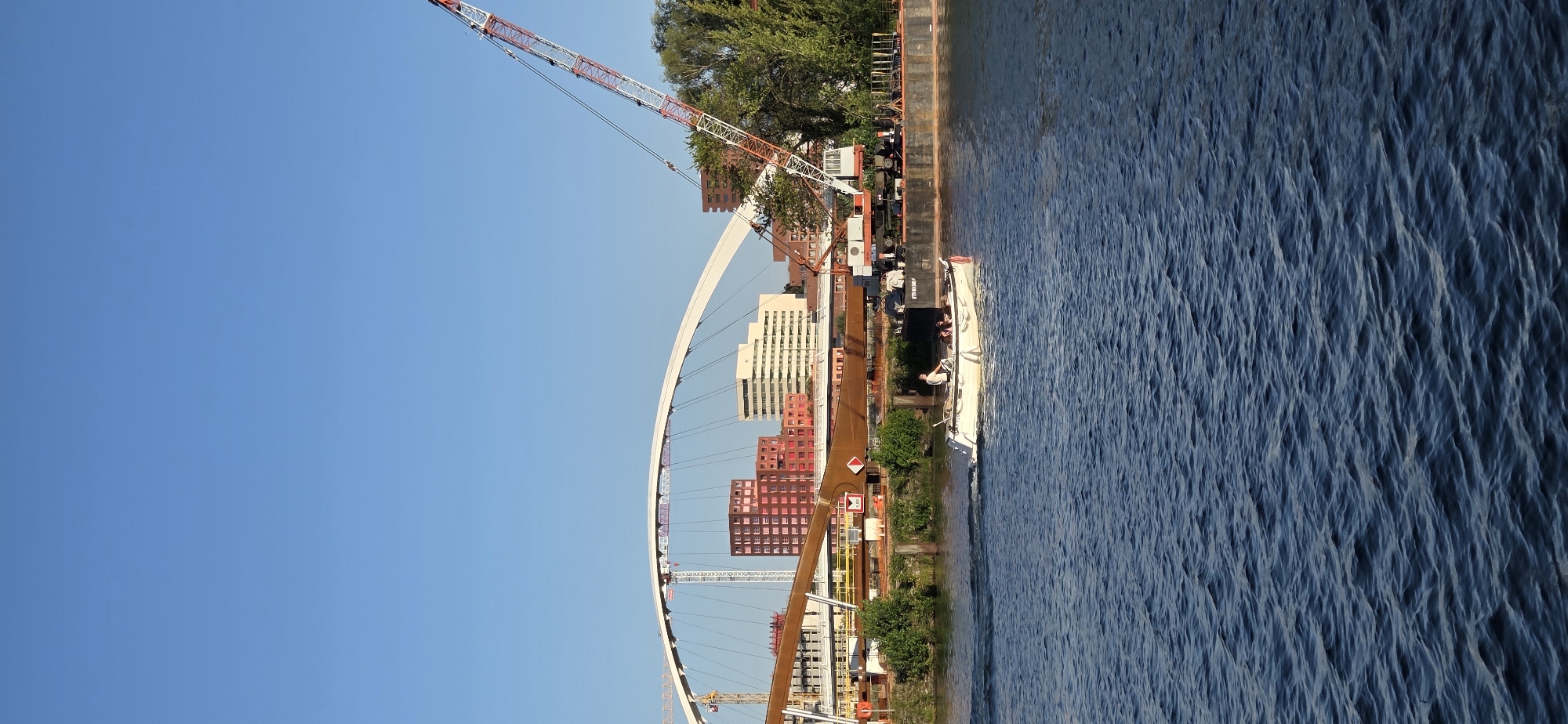 A wide shot shows a body of water with ripples under a clear blue sky. A white motorized boat with four people on board is moving from left to right across the water. The boat has a single person steering at the back and three passengers further forward.
In the background, a construction site is visible with a large white arched bridge under construction. A tall, red and white crane is positioned to the right of the bridge. Several red-brick buildings and a white building with many windows are behind the bridge, suggesting an urban environment. The scene appears to be along a river or canal in Strasbourg, France, given the architectural style and the presence of a large river crane and extensive bridge construction, common in port cities undergoing infrastructure development. The water appears calm except for the wake of the boat.