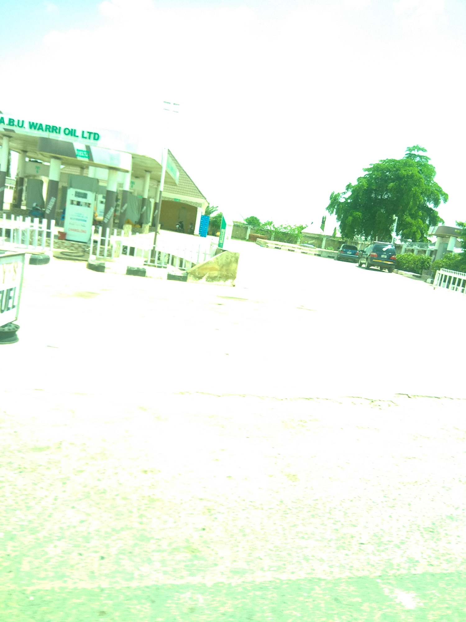 The image depicts a scene at a gas station. A sign overhead reads "A.B.U. WARRI OIL LTD". Two fuel pumps are visible, one with a white structure and a blue digital display, and another partially obscured. Several white bollards with black tires at their base line the area around the pumps. A sign with green lettering on a white background reads "FUEL". In the background, a building with a tiled roof appears to be part of the gas station. Further in the distance, two cars are parked on a road, and a large, lush green tree dominates the right side of the frame. The sky is bright and appears to be a clear, sunny day. The scene is set in Jalingo, Nigeria. There are no visible people in the image. The overall impression is of a typical, operational gas station under bright daylight.