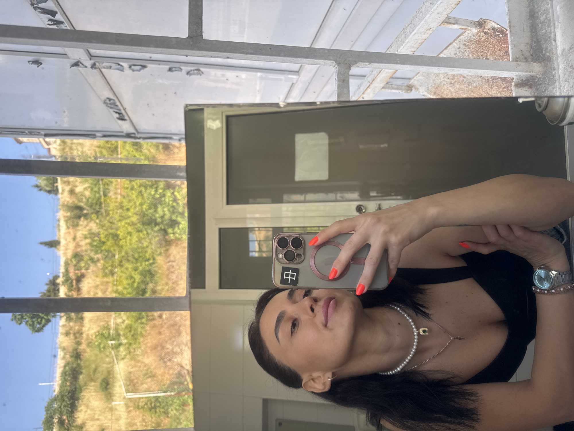 A young woman with dark hair is taking a mirror selfie in an urban outdoor setting, possibly a balcony or rooftop in Tbilisi, Georgia. She is wearing a black top, a pearl necklace, a silver necklace with a green pendant, and a silver watch on her left wrist. Her fingernails are painted bright orange. She is holding a smartphone in her right hand, with a gray case and a circular mirror attachment. The background reveals a sunlit hillside with dry vegetation and scattered green trees. A building with arched windows is visible on the hill. To the right, a white metallic structure with rusted elements suggests scaffolding or an industrial element. The reflection in the mirror also shows a door with a frosted panel.