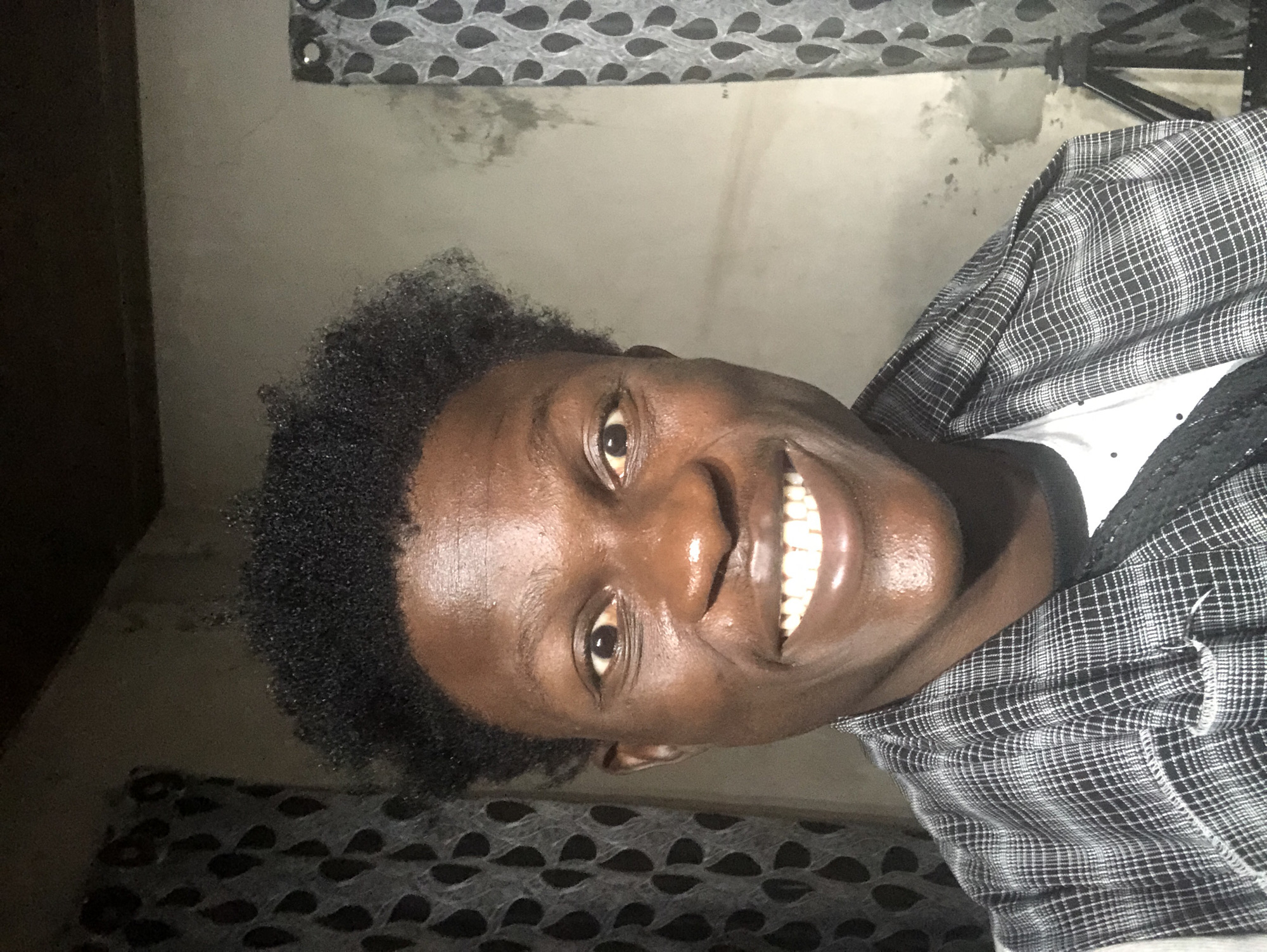A selfie of a young Black man is taken indoors, possibly in Ajah, Nigeria, as indicated by the context. He is positioned centrally, looking directly at the camera with a wide, genuine smile, showcasing his teeth. The man has dark, curly hair and dark brown skin. He is wearing a black and white plaid shirt over a white t-shirt. The background consists of a gray, textured wall with some staining, and to his left and right, patterned curtains are visible. The lighting suggests an indoor setting, possibly at night or in a dimly lit room, with light directed from the front, highlighting his facial features and creating a slight sheen on his skin. The overall scene is simple and personal, focusing on the subject's expression.