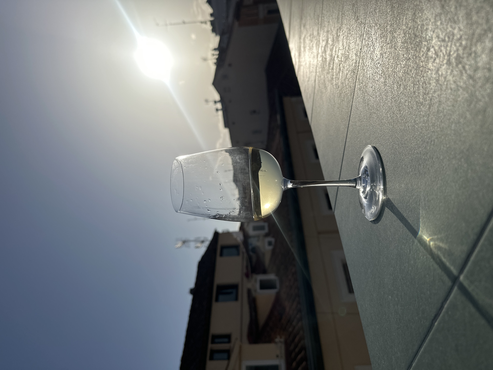 A low-angle shot depicts a wine glass on a gray tiled surface with a building, sky, and sun in the background. The wine glass is mostly empty, containing a small amount of a yellowish liquid. The base of the glass casts a distinct shadow on the tiles. The building in the background appears to be an older structure, with windows and possibly air conditioning units visible. The sun is bright and high in the sky, creating lens flare in the image. There is nothing to support the location of the image being in Madrid, Spain.