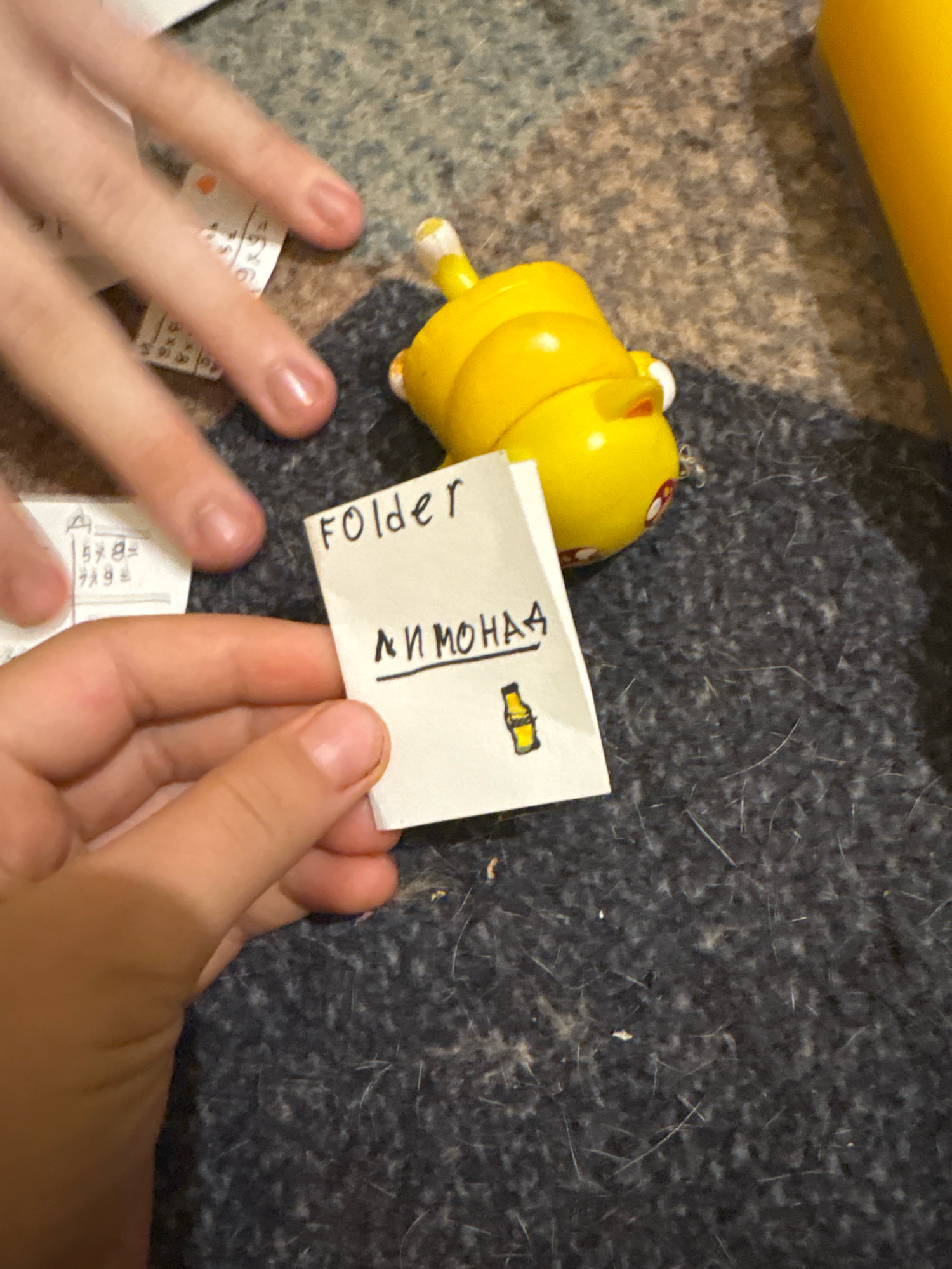 A close-up, high-angle shot captures a child's hands holding a small, folded piece of paper on a dark, textured surface. The paper has the word "Folder" written in English on the top, and below it, in Cyrillic script, it reads "лимона", meaning "lemonade". A small drawing of a bottle of lemonade is also on the paper. To the right of the paper, a bright yellow toy with red and white accents is visible. In the background, a faint glimpse of a yellow object can be seen. On the left side of the frame, a portion of a child's hand is visible, with faint writing and numbers on a white surface. The overall scene suggests an indoor, informal setting, possibly a home or classroom, with no discernible connection to Ashvem, India.