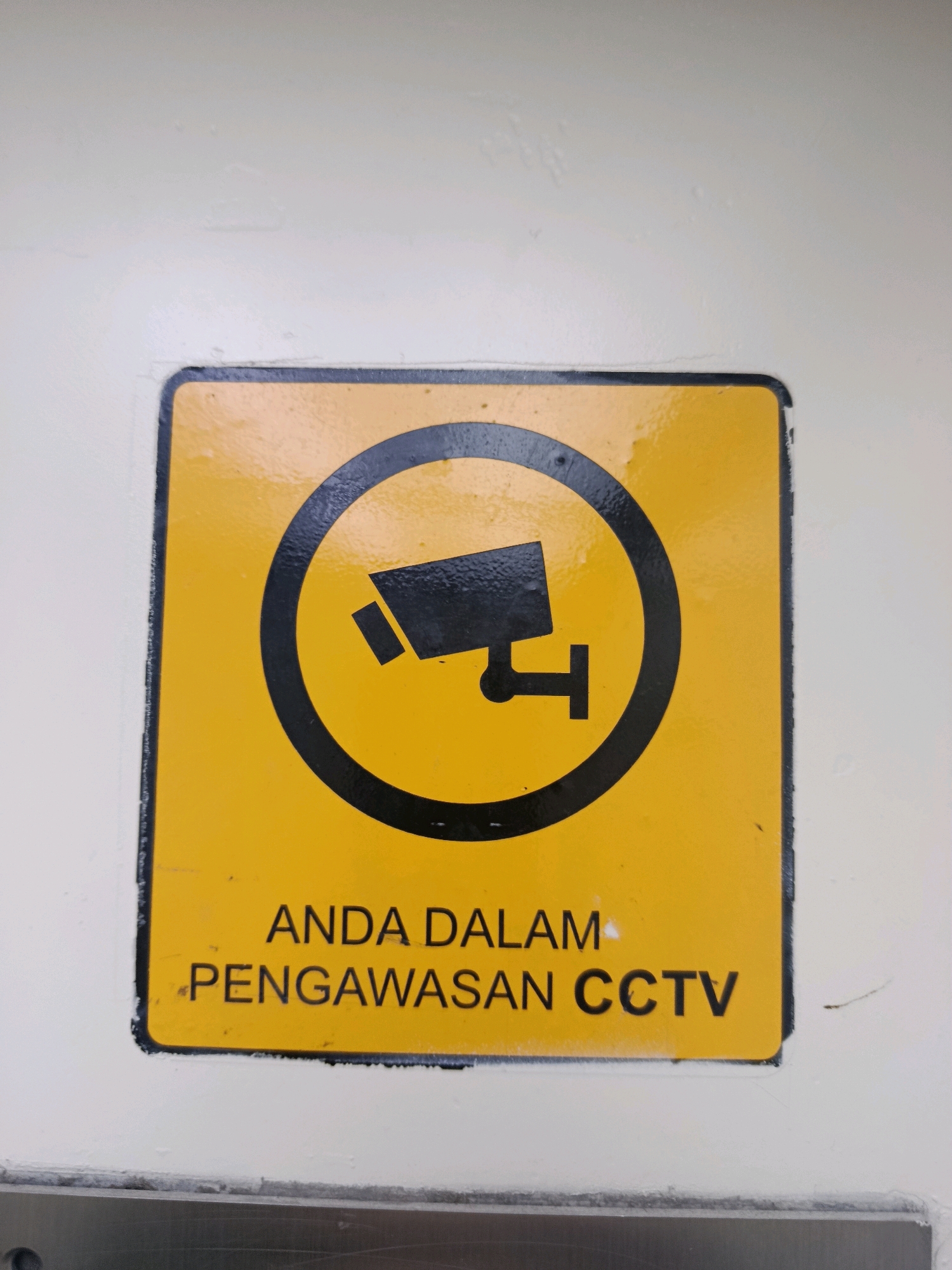 A yellow square sign with black border and symbols is mounted on a textured white wall. The sign depicts a black icon of a security camera within a black circle. Below the camera icon, text in black reads "ANDA DALAM PENGAWASAN CCTV". This translates from Indonesian to English as "YOU ARE UNDER CCTV SURVEILLANCE". The sign is likely located in South Jakarta, Indonesia, indicating a monitored area.