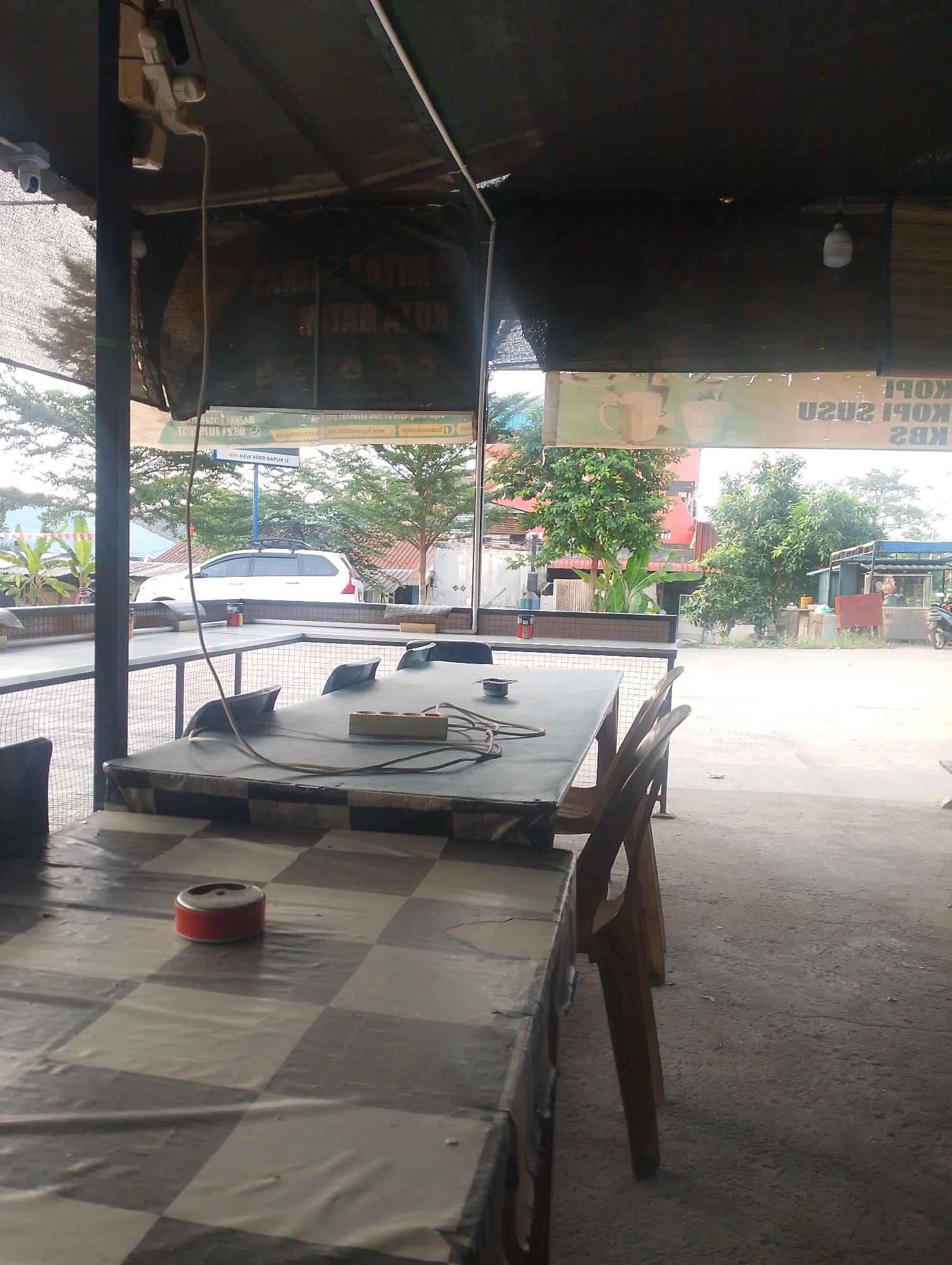 The image captures an outdoor seating area, likely a cafe or restaurant, in Batam, Indonesia. In the foreground, a checkered-patterned table is covered with a plastic tablecloth. On this table, an ashtray is placed. To the right of this table, several wooden chairs are visible, one of which is angled towards the viewer.
Behind this seating area, another table is set with electrical cords and an extension cord with multiple outlets. A white SUV with a roof rack is parked outside the establishment, with trees and a street visible in the background. The overall ambiance suggests a casual, open-air dining or resting spot. The establishment displays signage, though much of it is obscured or not clearly legible in the provided image. One visible sign has text in Indonesian, and the overall visual cues suggest it is a place that serves beverages like coffee. The lighting indicates it is daytime, with the sun casting shadows.
